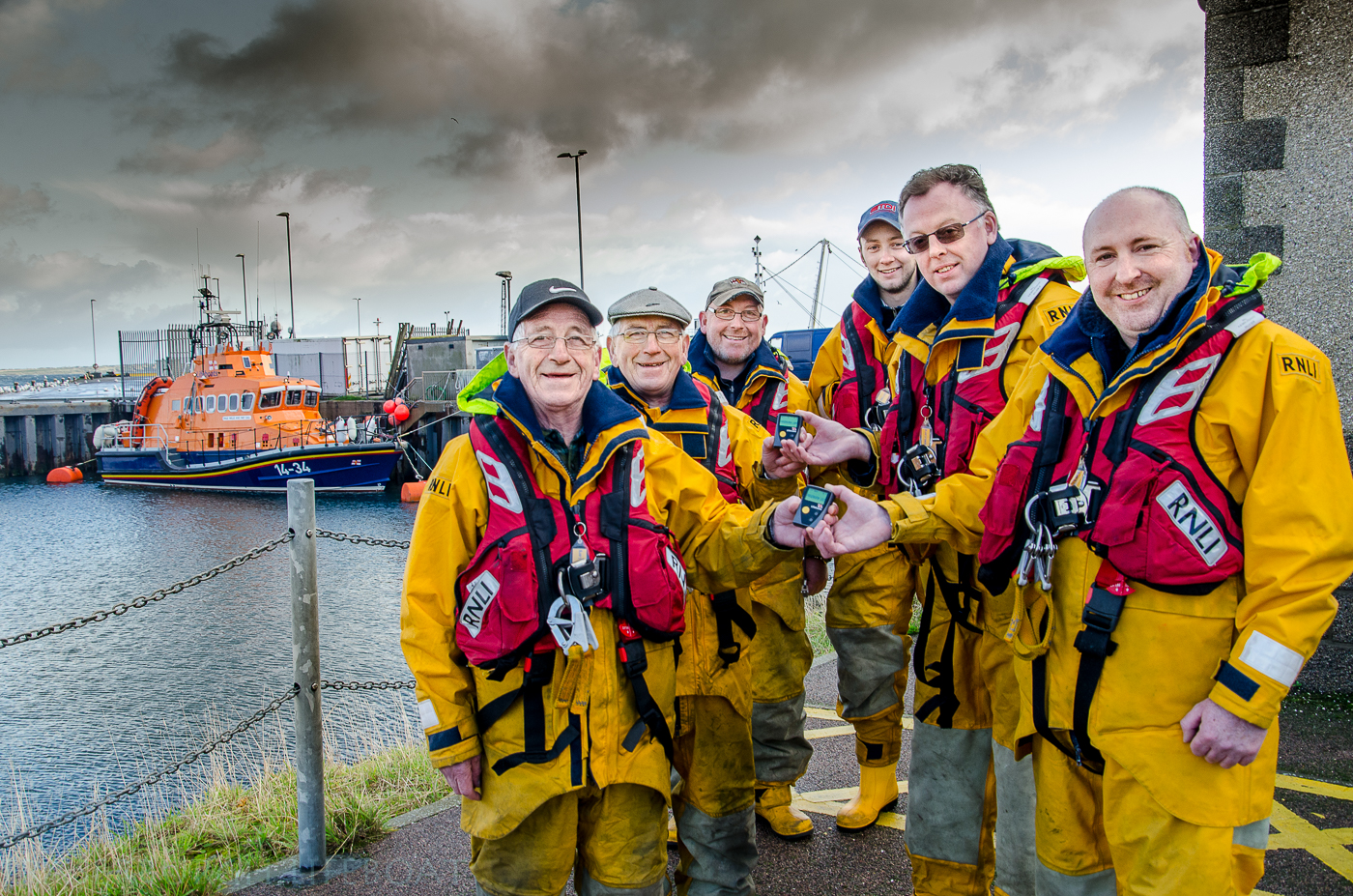 A group photo taken outside the Fraserburgh Lifeboat Station with the Fraserburgh Lifeboat the Willie and May Gall in the backgound. Twin Brothers Victor senior and Albert Sutherland hand over their LOM and DLA pagers to their successors Hamish Partridge and Stuart Fenty. Looking on are coxswain Vic Sutherland and volunteer mechanic Shane Richardson