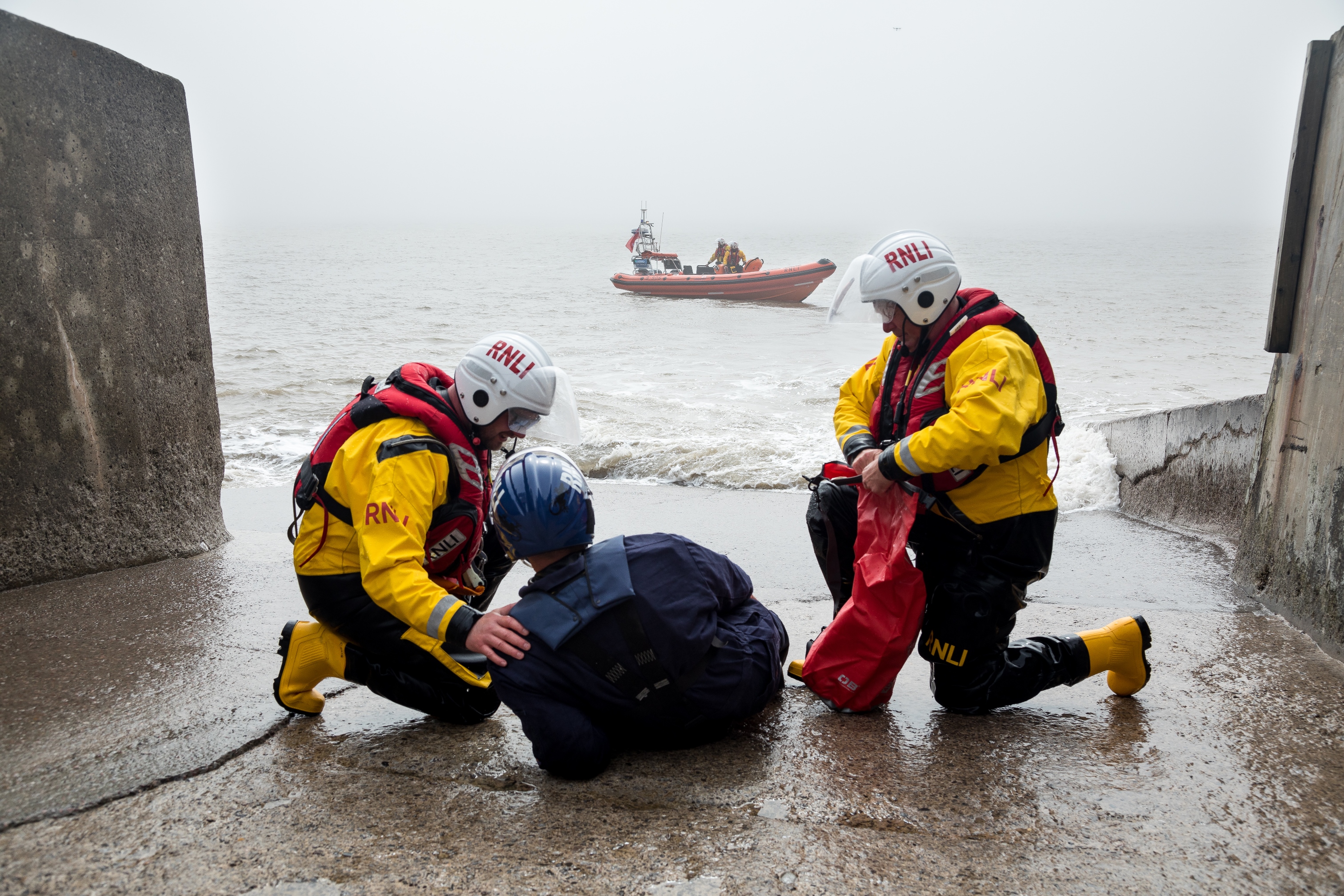 Two crew members on the shoreline giving casualty care to a man, with an inshore lifeboat in the background