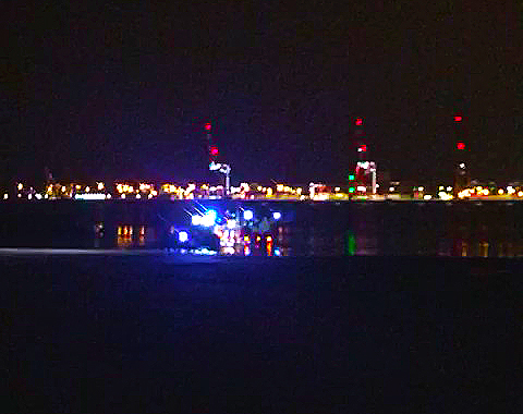 In darkness, lifeboat and trailer lit up in water in front of a back drop of Liverpool port