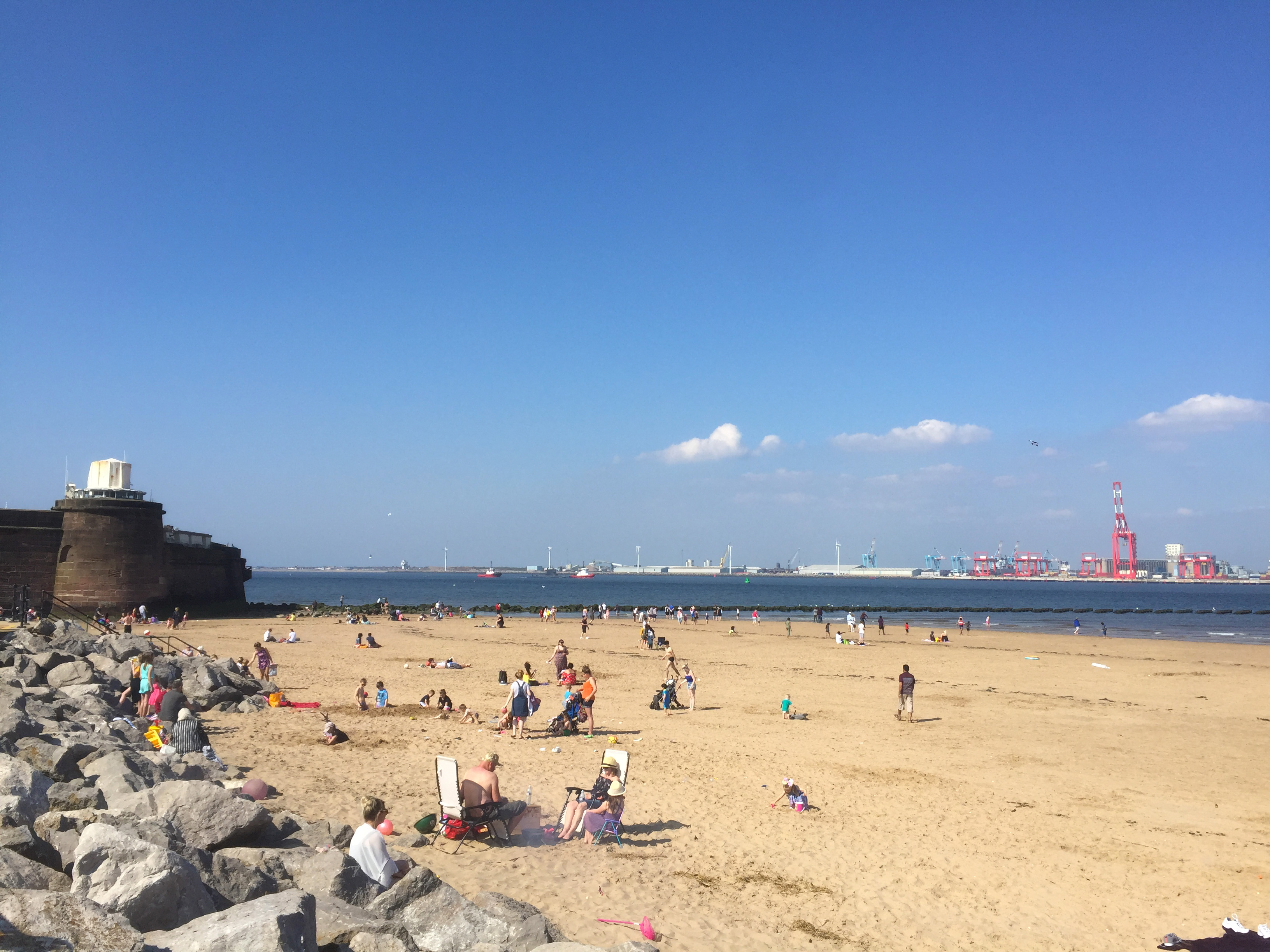 Blue sky overhead with Fort Perch Rock in the distance,rocks on the left and to the right an expanse of sand busy with people and a glimpse of the sea