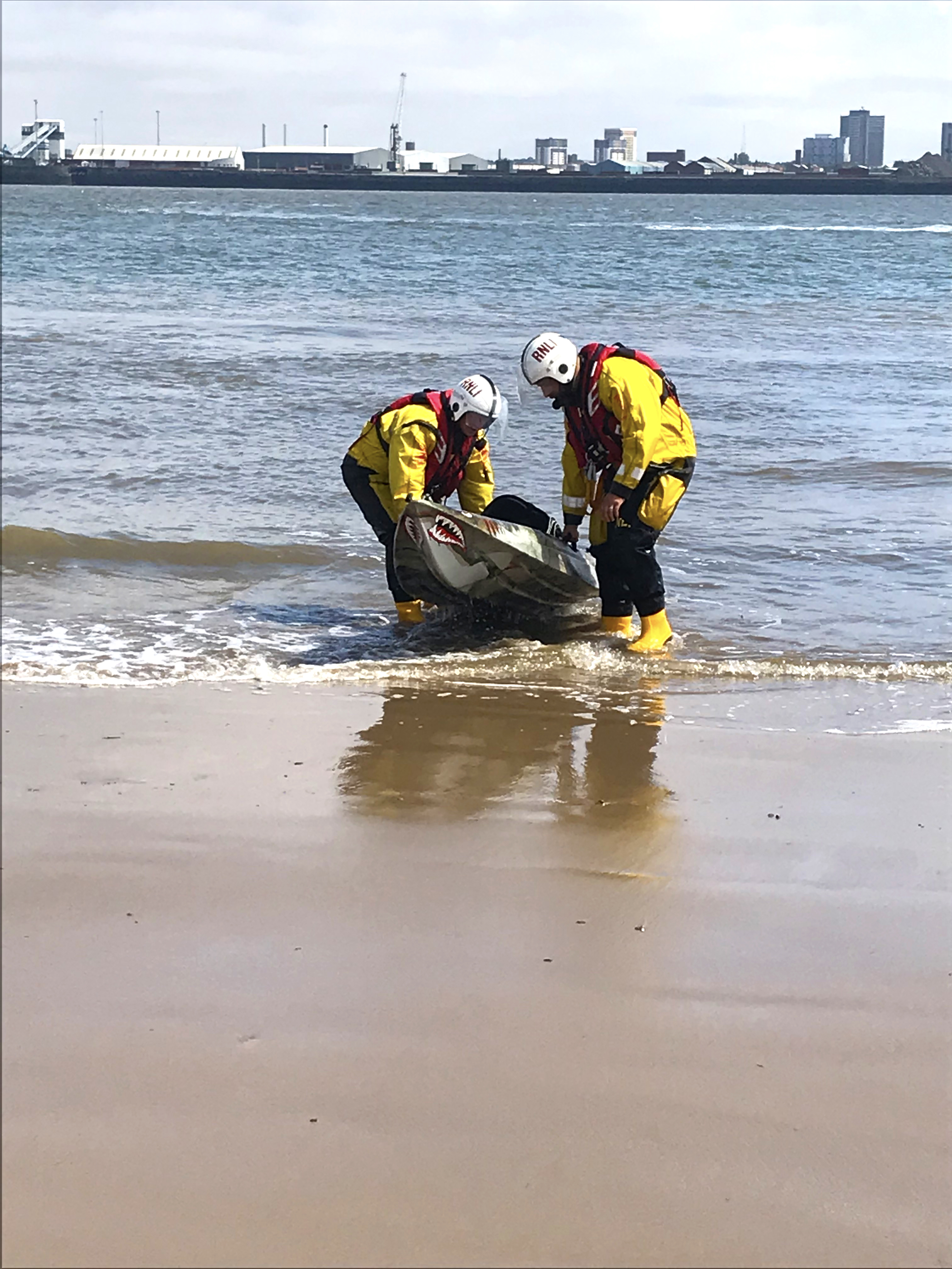 Foreground of sand then two RNLI shore crew either side of the kayak in the shallows as they start to exit the water.