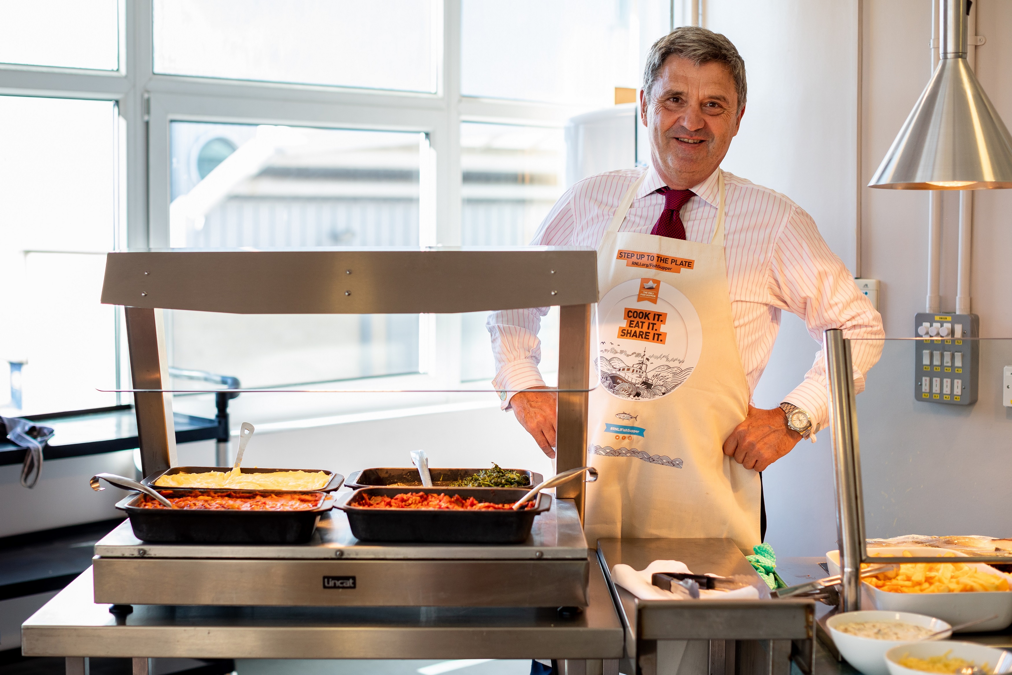 RNLI Chief Executive Paul Bossier is stood behind a serving counter in the staff canteen wearing a Fish Supper apron