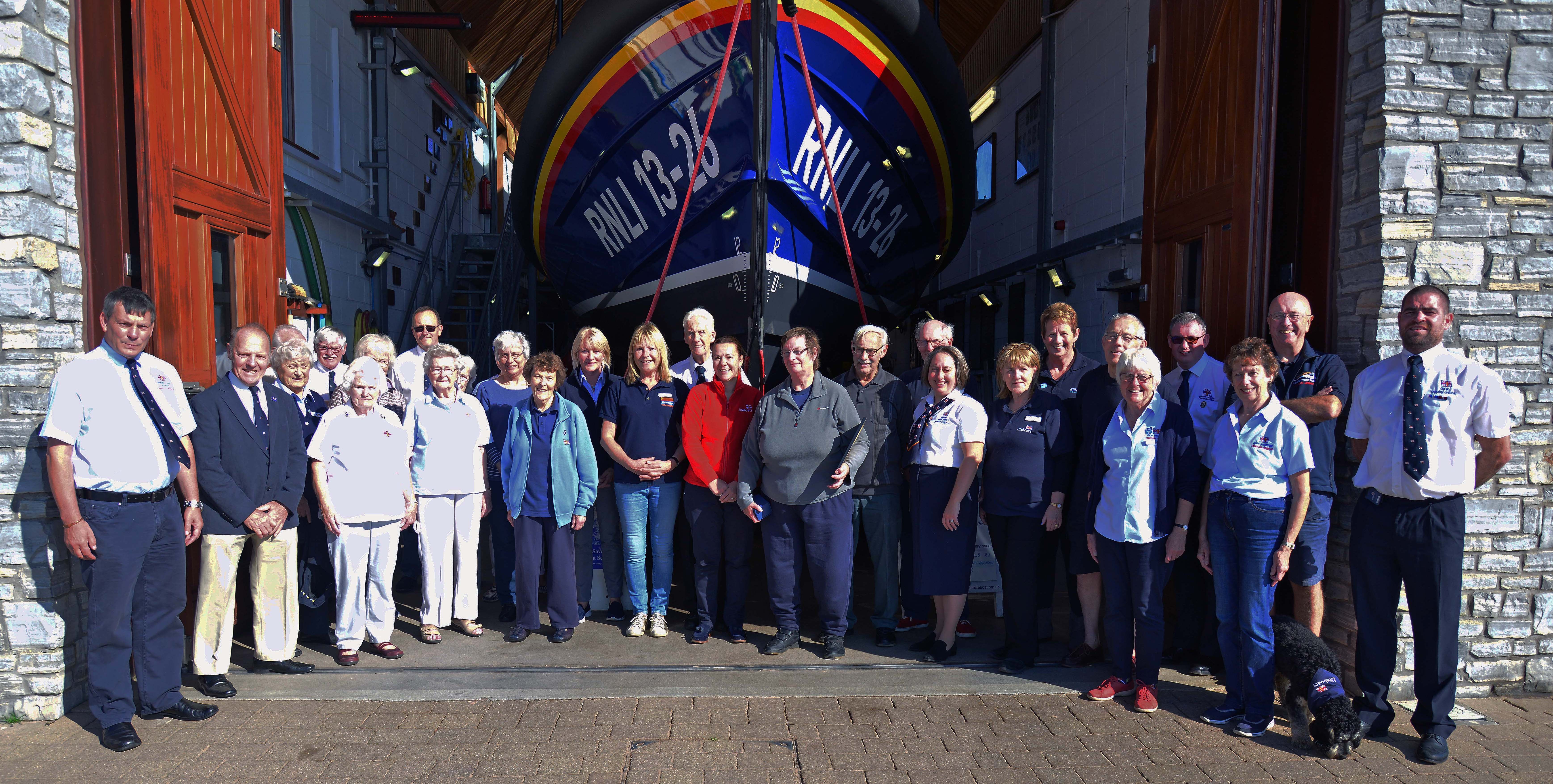 Volunteers from Exmouth RNLI shop, fundraising, Lifeboat Management Group and station stand in front of the lifeboat station with Bernice Williams and her husband, Geoffrey