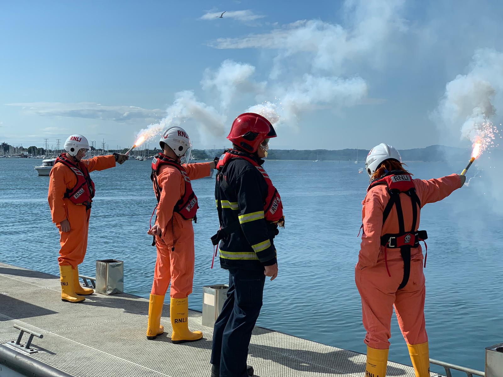 Three lifeboat crew-members hold lit emergency flares at arm's length while and instructor looks on