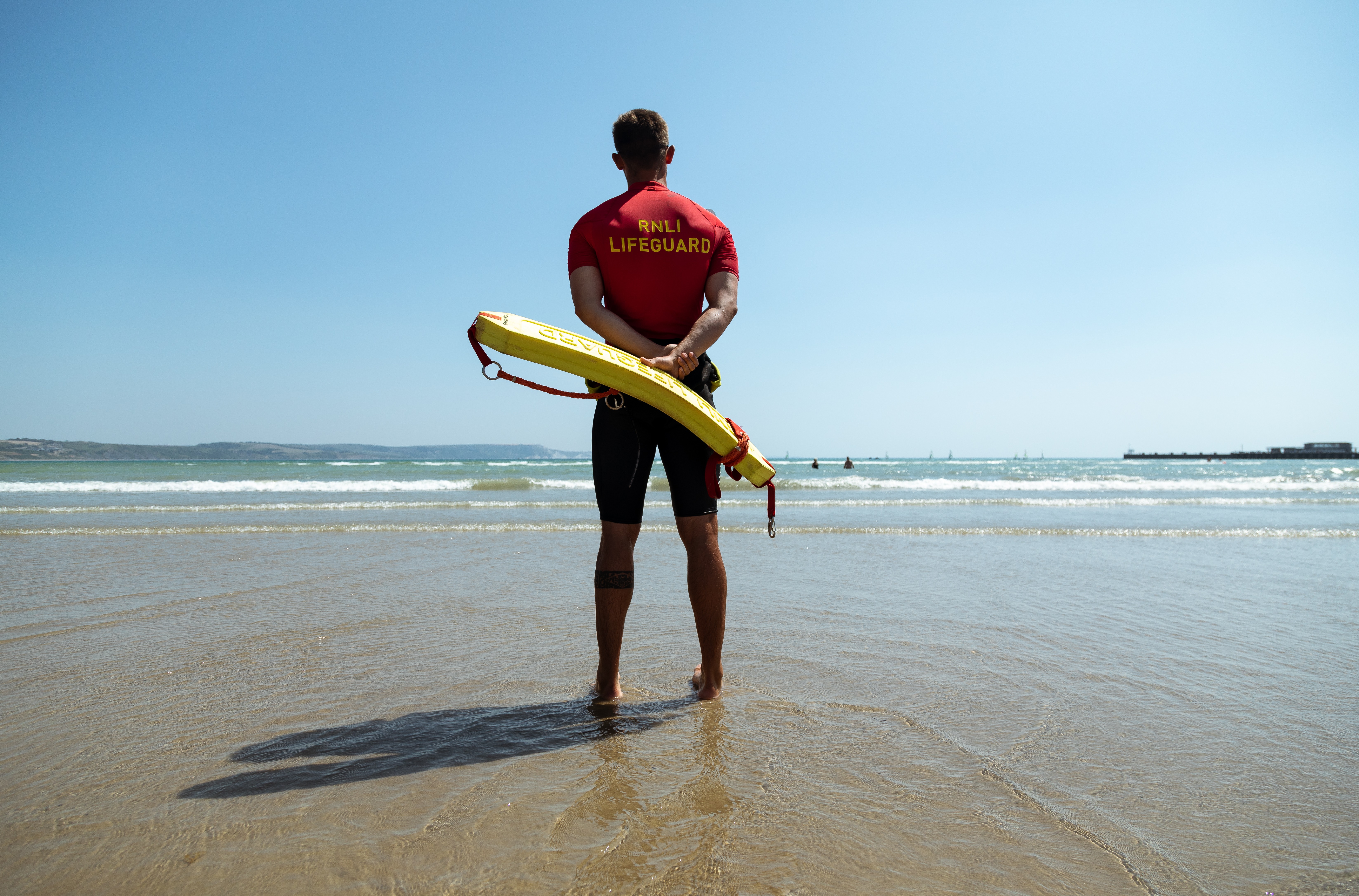 Photo of the back of lifeguard on the shoreline and monitoring the sea holding a yellow rescue tube.