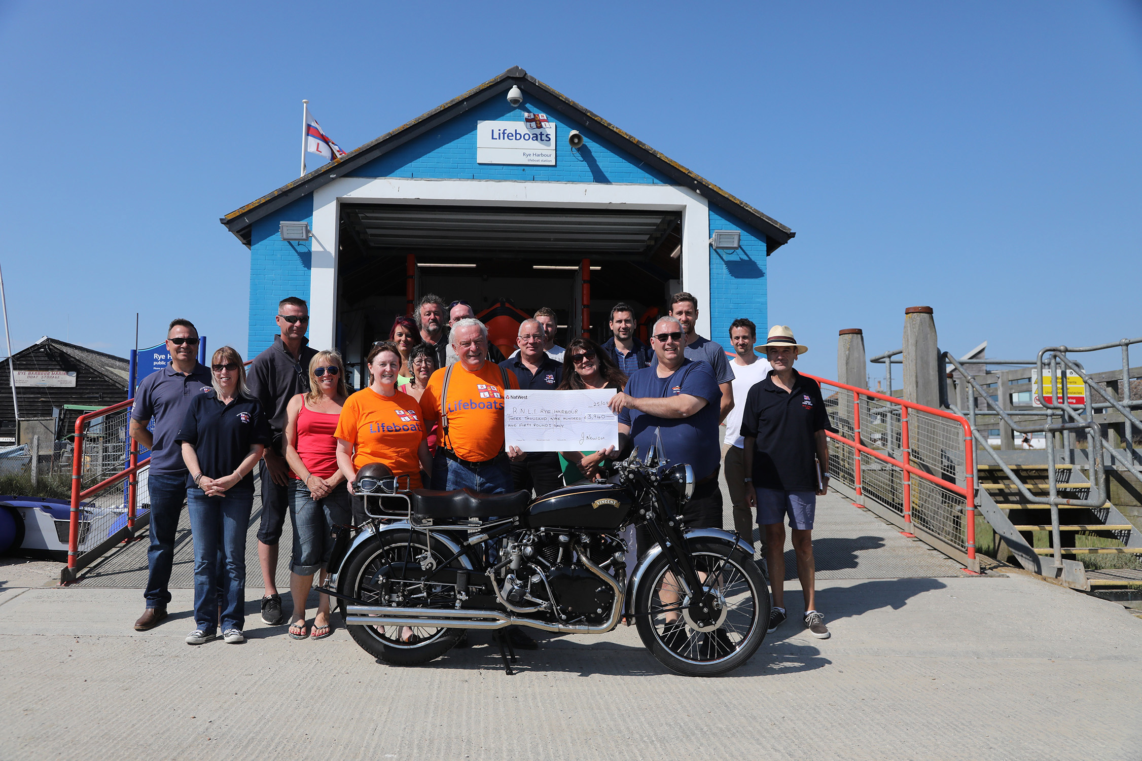 There is a crowd of RNLI crew and well-wishers in front of the boathouse. John Newson proudly holds the cheque for the money he has raised