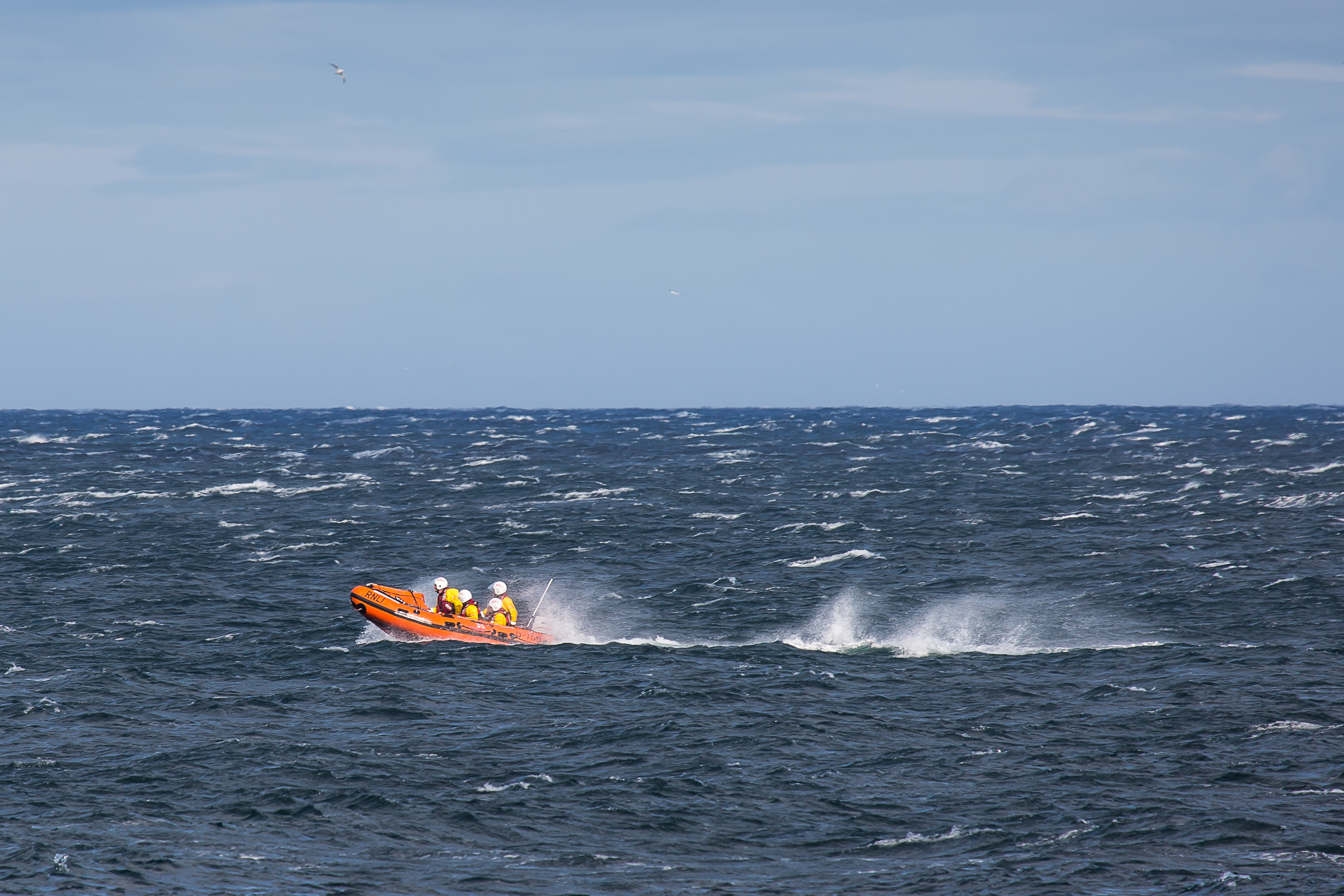 Dunbar ILB races through rough conditions to locate casualty. Blue skies overhead and excellent visibility. The azure blue sea is rough and capped with many white horses. The bright orange boat with it's bow risen in the surf travels from right to left and is followed by a heavy wake that is broken by the strong wind creating a lot of spray. 4 crew members are visible on board.