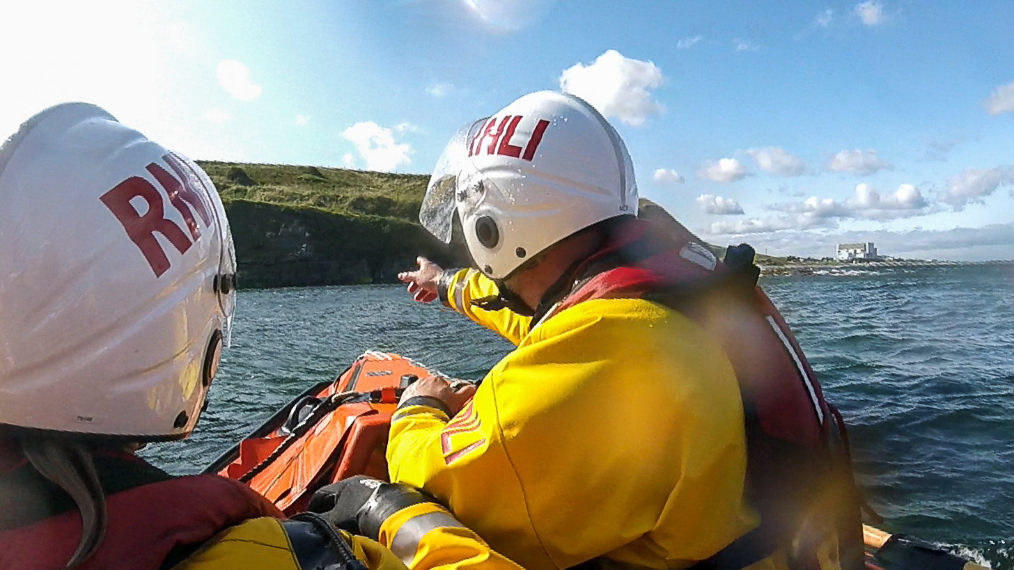 Two crew members sit at the bow of the ILB. The crew member on the right has his arm raised and is pointing at the shoreline. In the distance, beyond the white helmets and yellow suits of the crew, the shore is visibly rough and ragged with low cliffs rising straight from the sea. Torness power station is visible in the distance on the far right. The sky is blue and dotted with small cumulonimbus clouds.