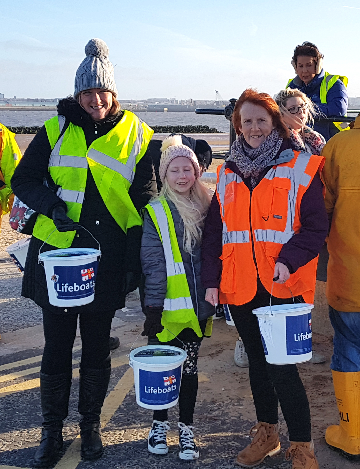 Two women stand with a younger child between them, with all three holding RNLI collection buckets
