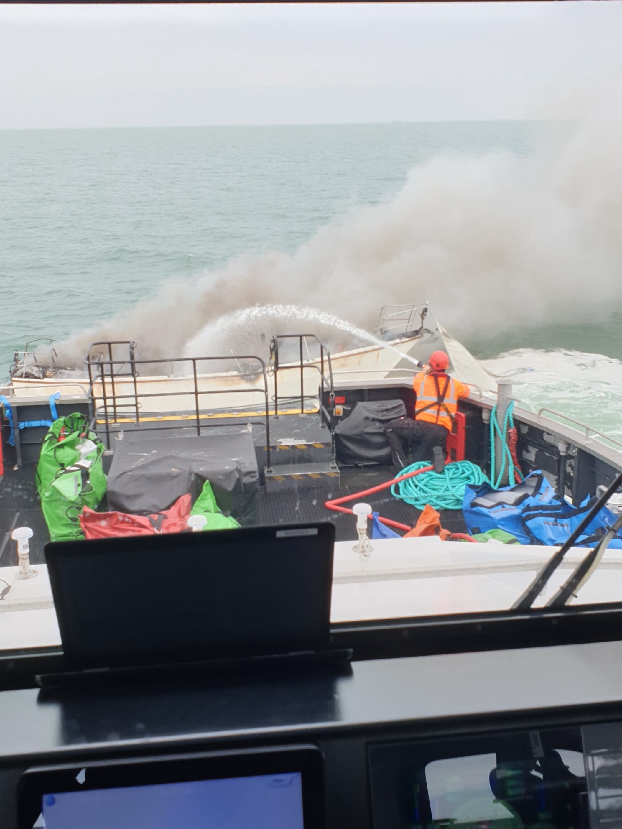 image taken through the windscreen of the windfarm vessel of the smoke and a crew member hosing the burning yacht.