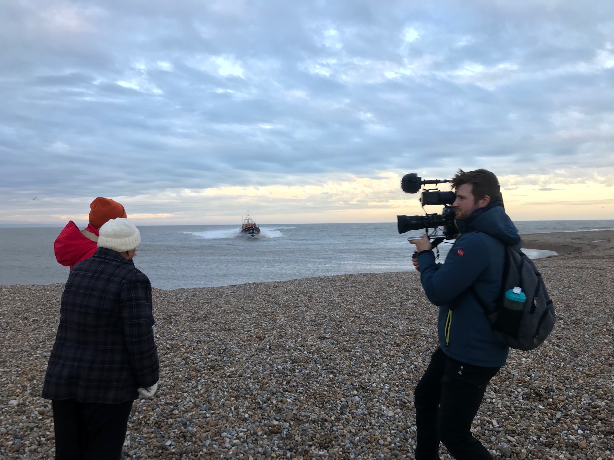 Two ladies stand watching the lifeboat which is heading towards the beach. A cameraman films them