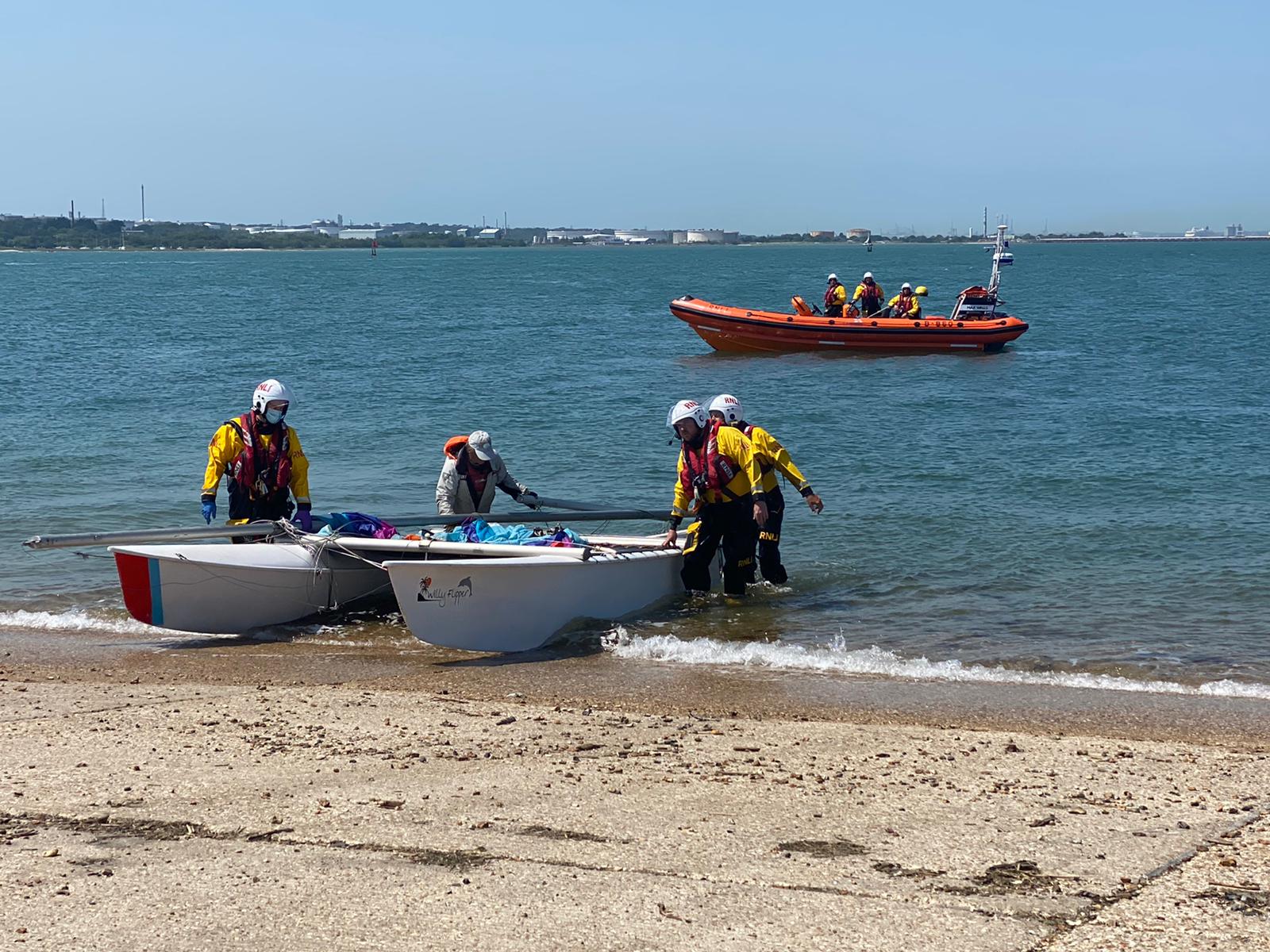 Photo showing the Calshot RNLI crew bringing the dismasted catamaran ashore with the lifeboat in the background.