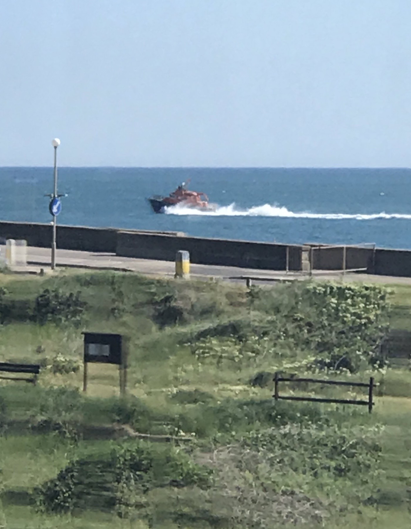 Viewed set back from the beach behind open grassland, the Severn Class lifeboat ‘David and Elizabeth Acland’ speeds through sea, close in to shore.