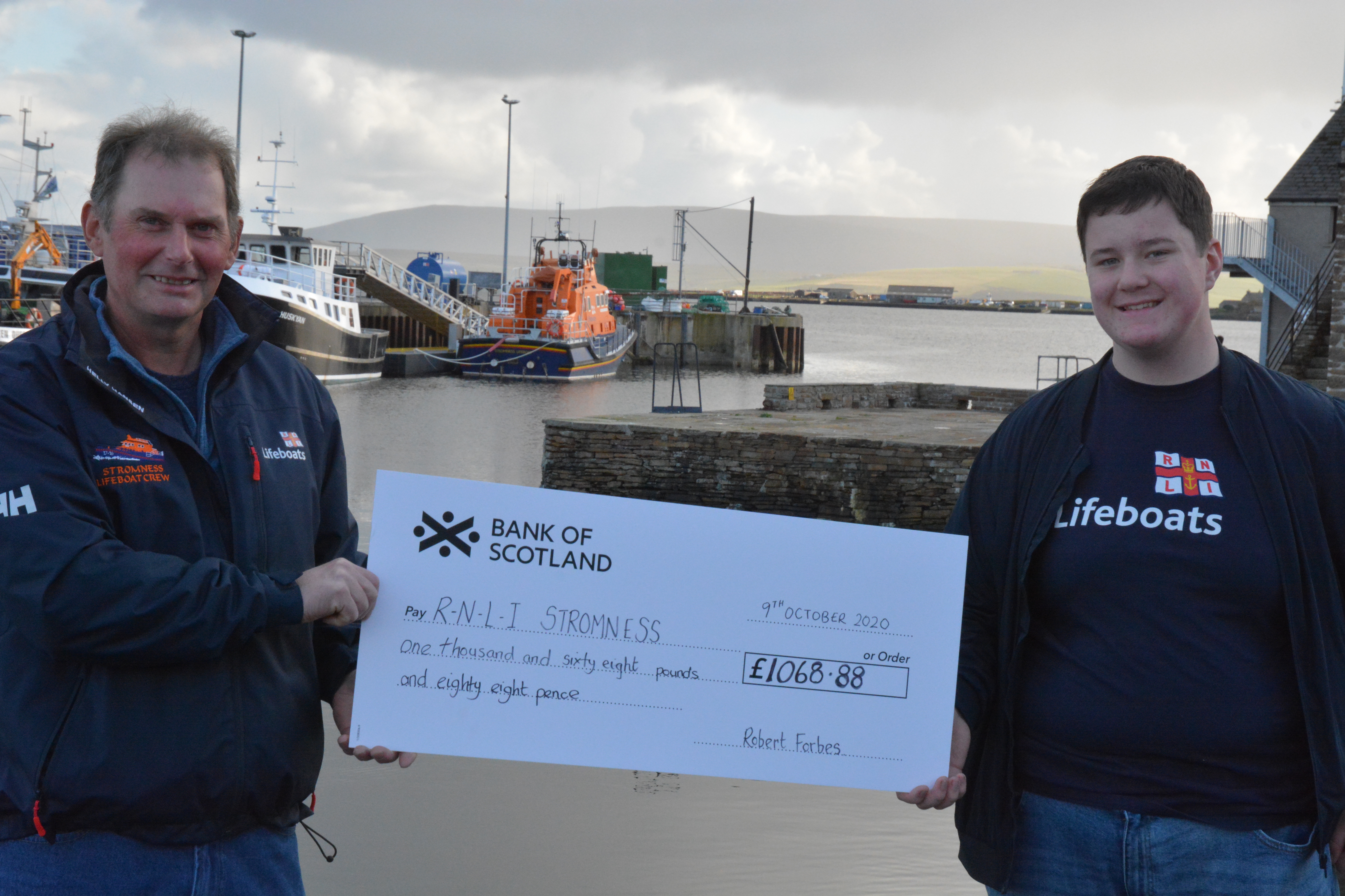 Robert Forbes (right) handing large cheque to Colin Mowat with Stromness lifeboat in the background