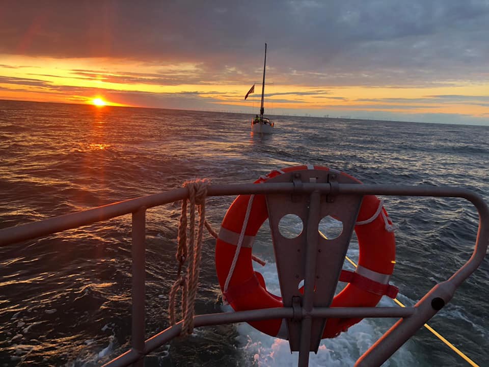 The stern rails of a lifeboat can be seen witha yacht in tow and the sunset behind