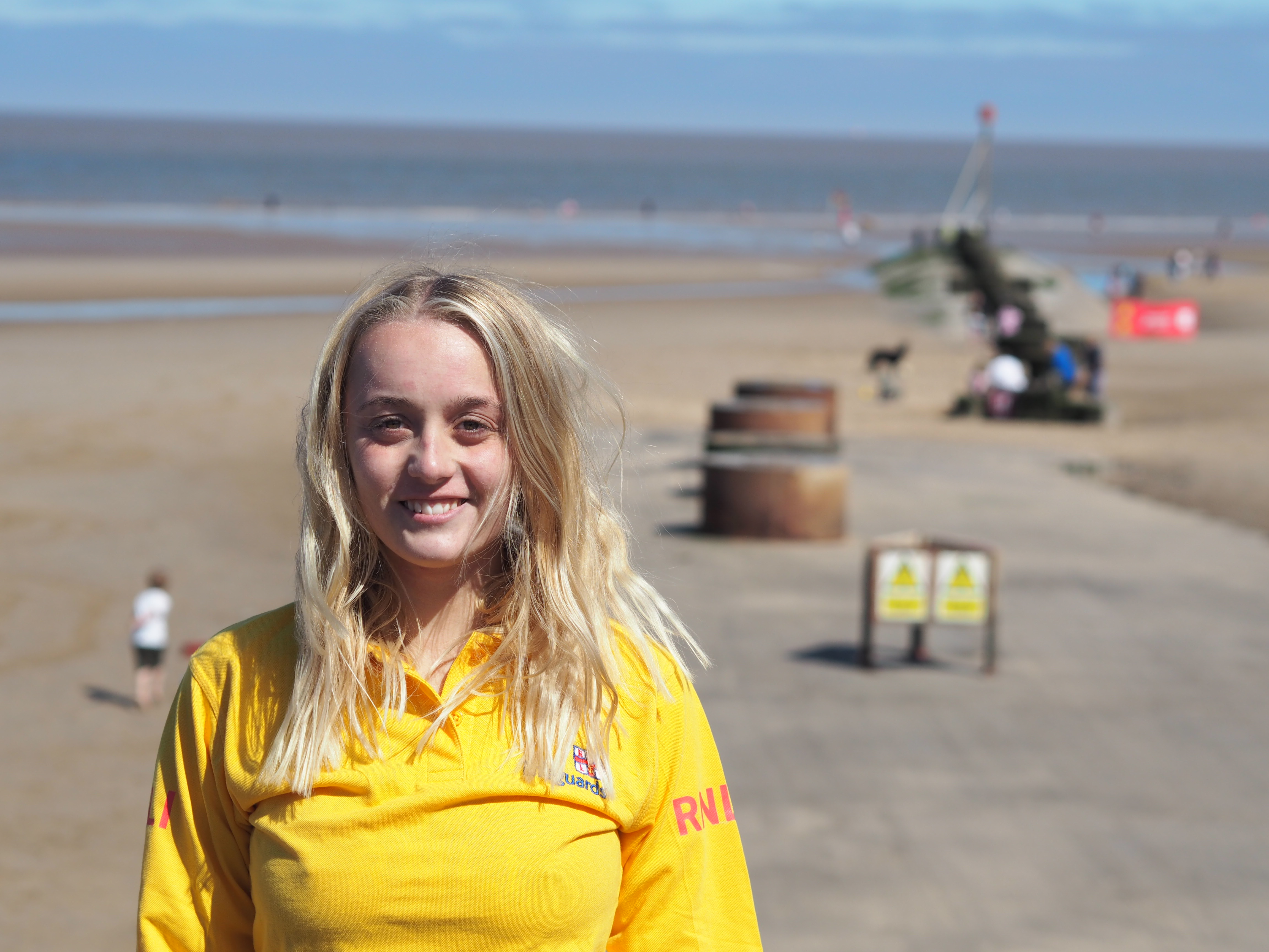 A young female lifeguard stands in front of the camera in a yellow RNLI lifeguard top. Behind, people are seen on a sandy beach with the sea in the distance.