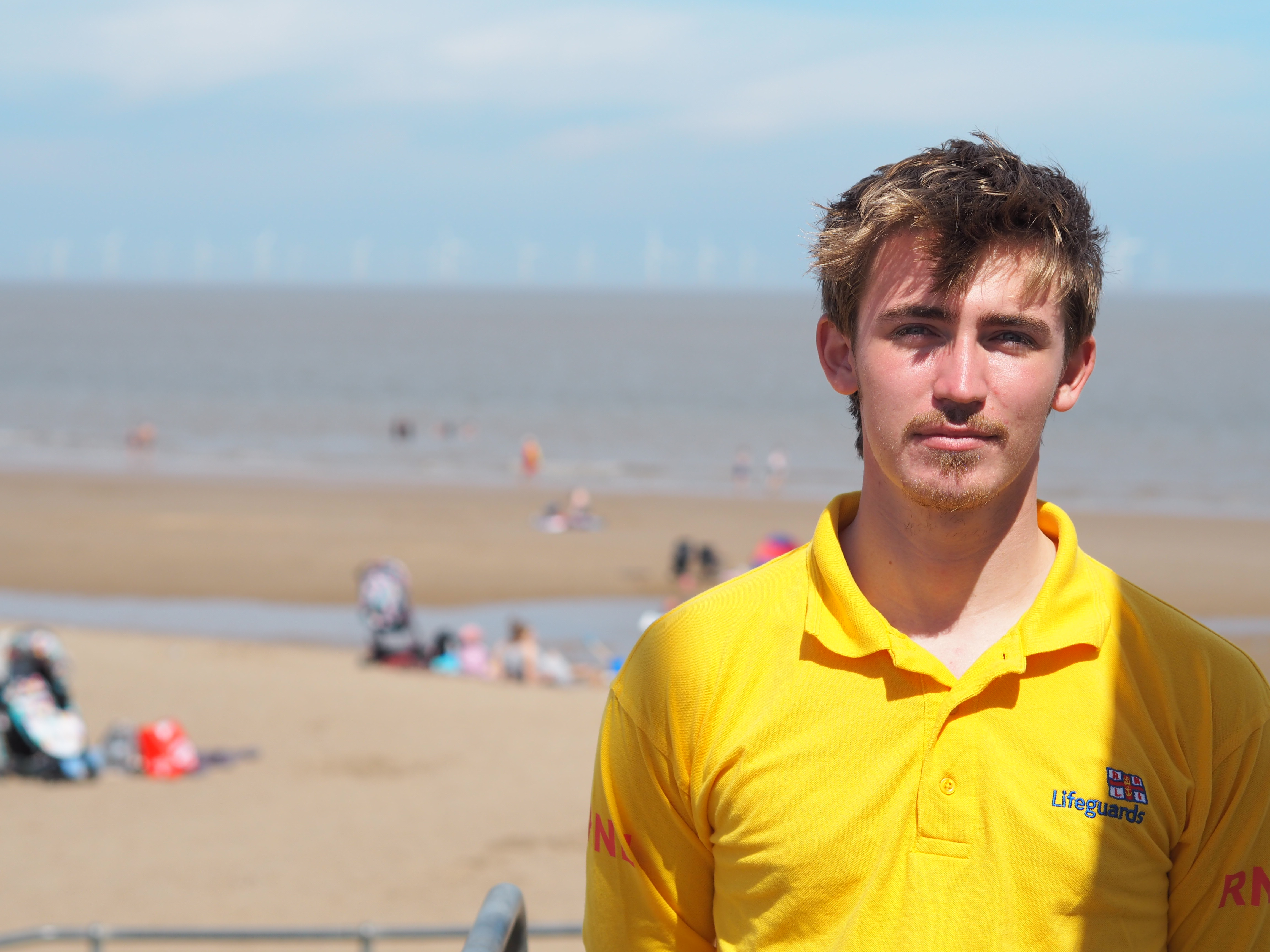A young male lifeguard stands in front of the camera in a yellow RNLI lifeguard top. Behind, people are seen on a sandy beach with the sea in the distance.