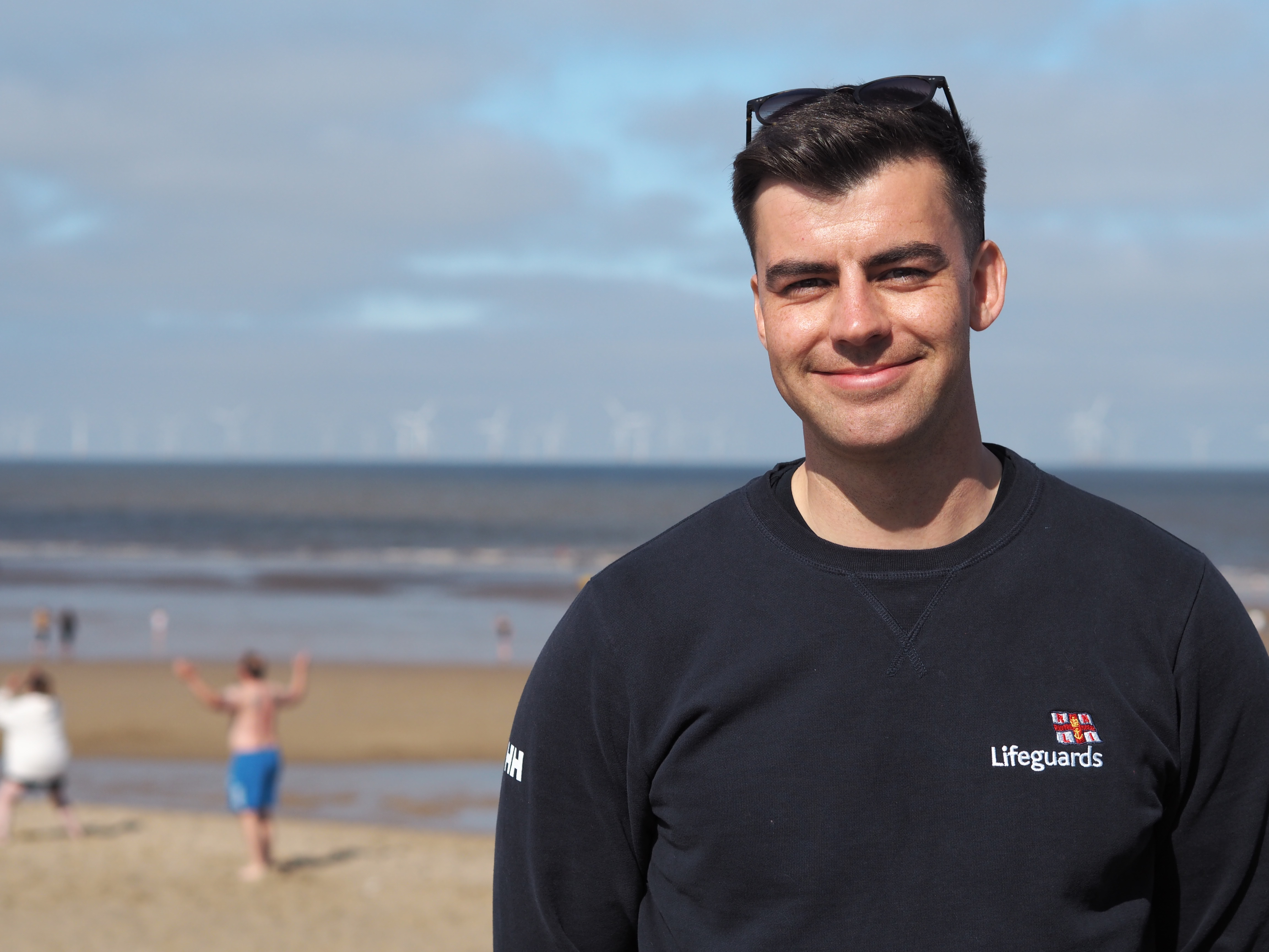 A young man in a navy jumper that says lifeguards on it and has the RNLI symbol stands in front of a sandy beach. Behind him, there is the sea.