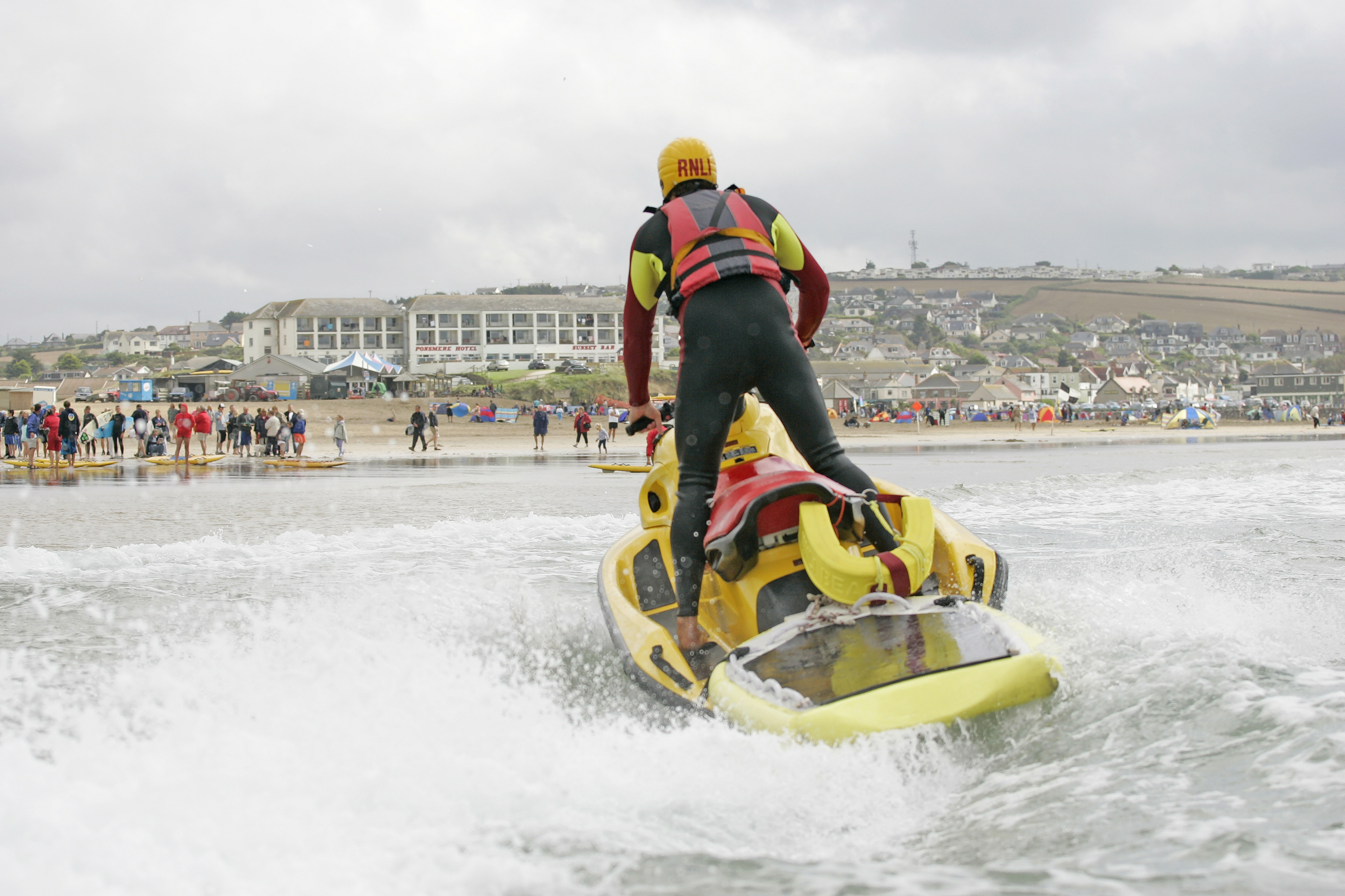 A person stands up on a yellow jet ski that has rescue equipment on the back. The person is in a wetsuit and wearing a yellow RNLI helmet and a red lifejacket.