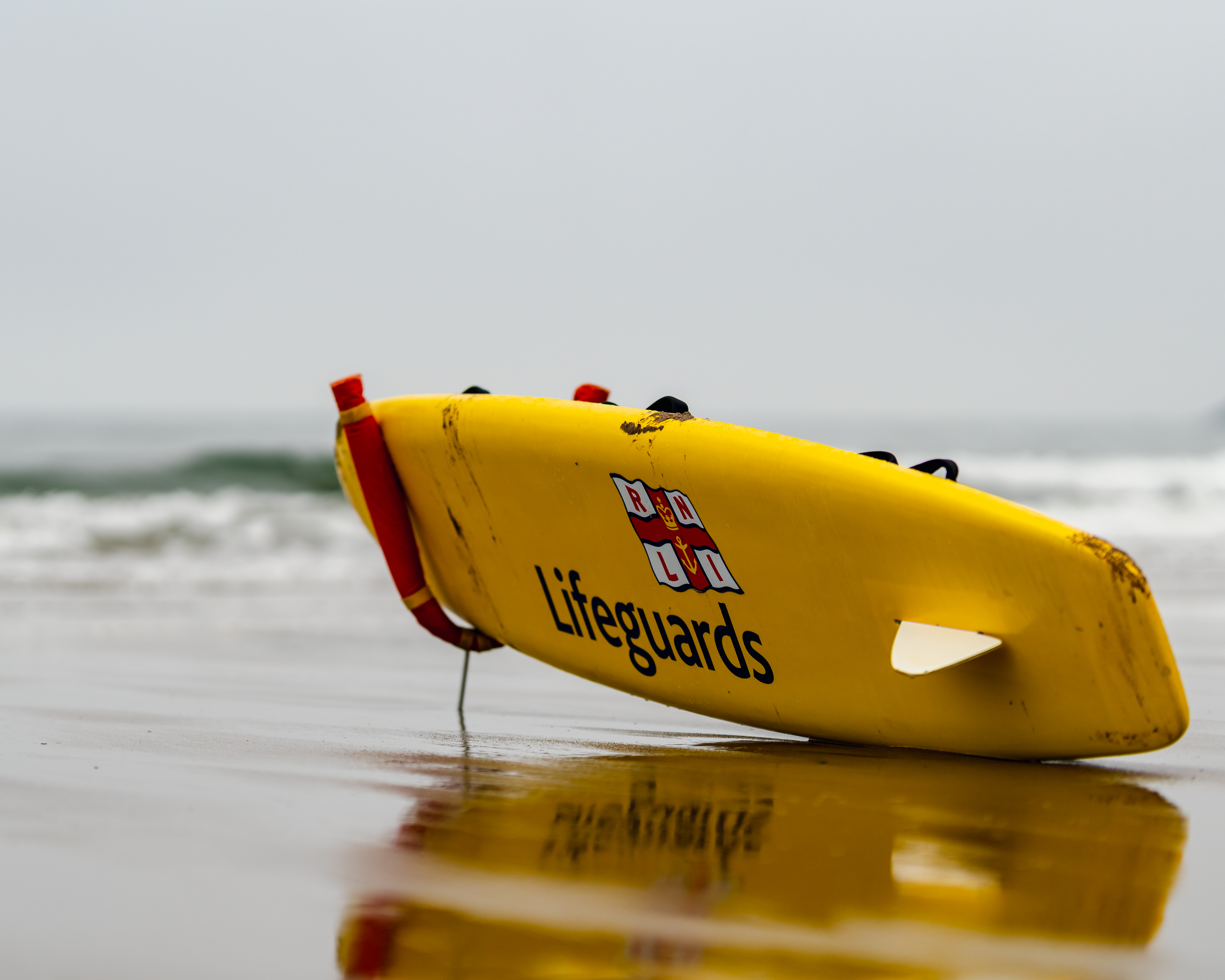 A yellow surf board lies on the sandy beach in front of the sea. On the side of it is the RNLI logo and the word lifeguards.