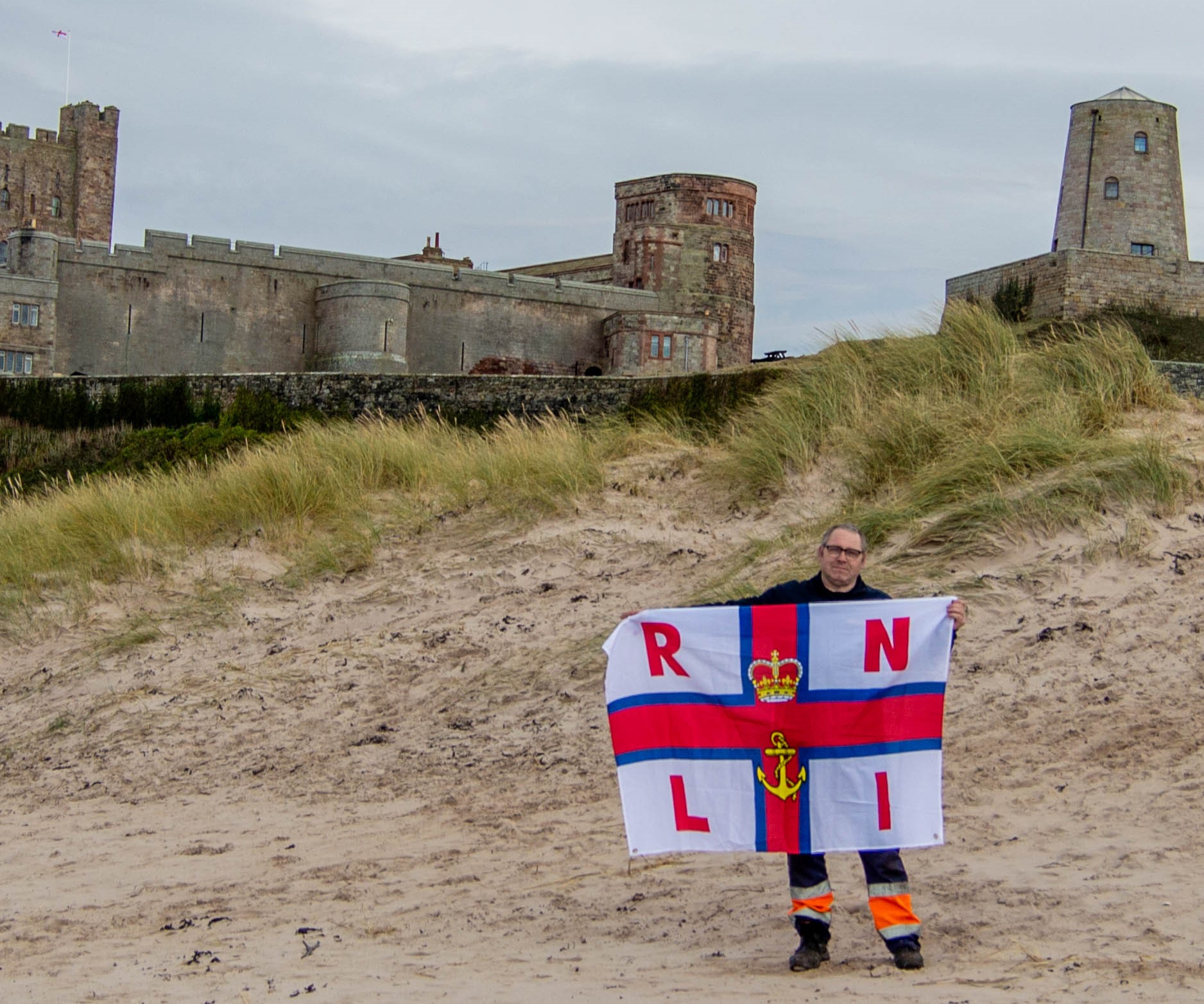 Chris Mason stands outside of Bamburgh Castle holding an RNLI flag