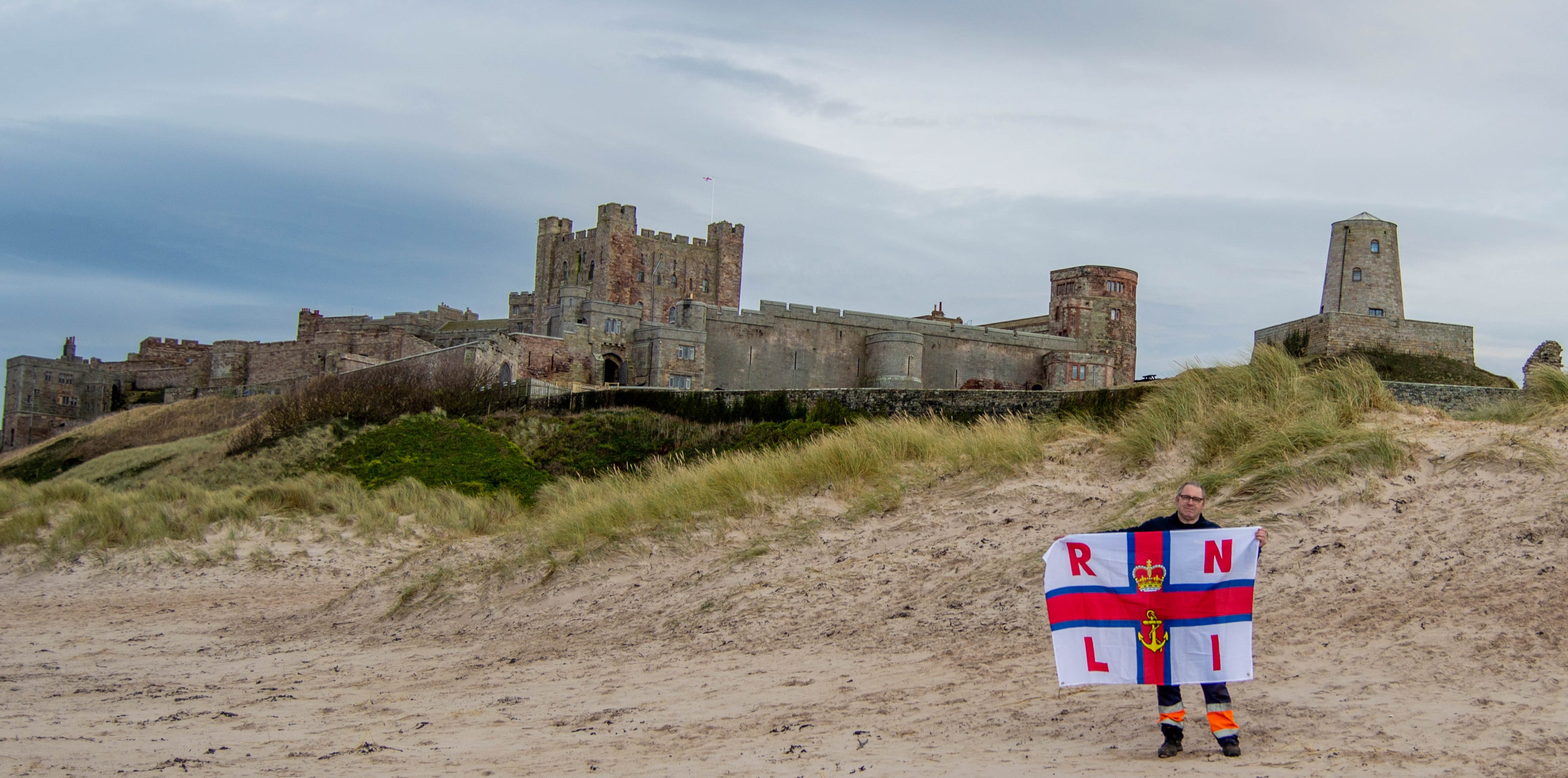 Chris Mason stands outside of Bamburgh Castle holding an RNLI flag