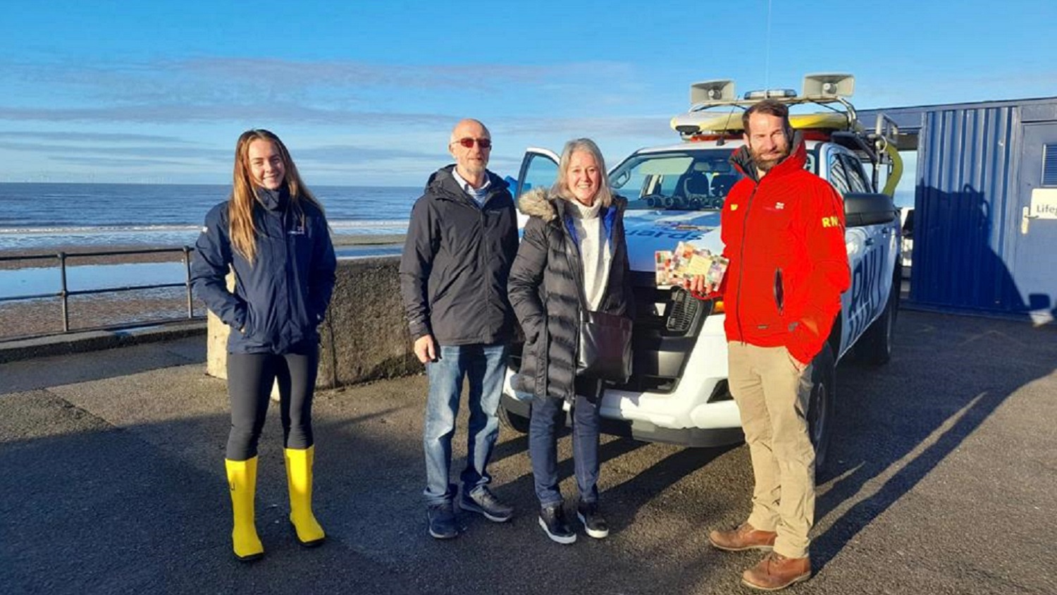 A group of four people, two women and two men, stand in front of a white RNLI rescue vehicle with the sea in the background.