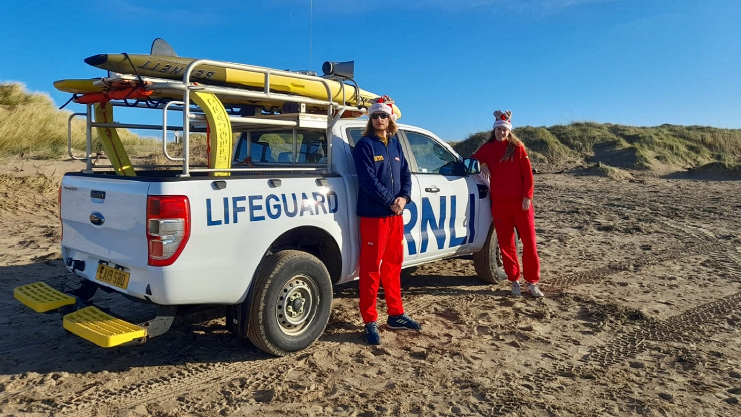 A female and male lifeguard stand in front of RNLI rescue vehicle in festive head wear on a beach.