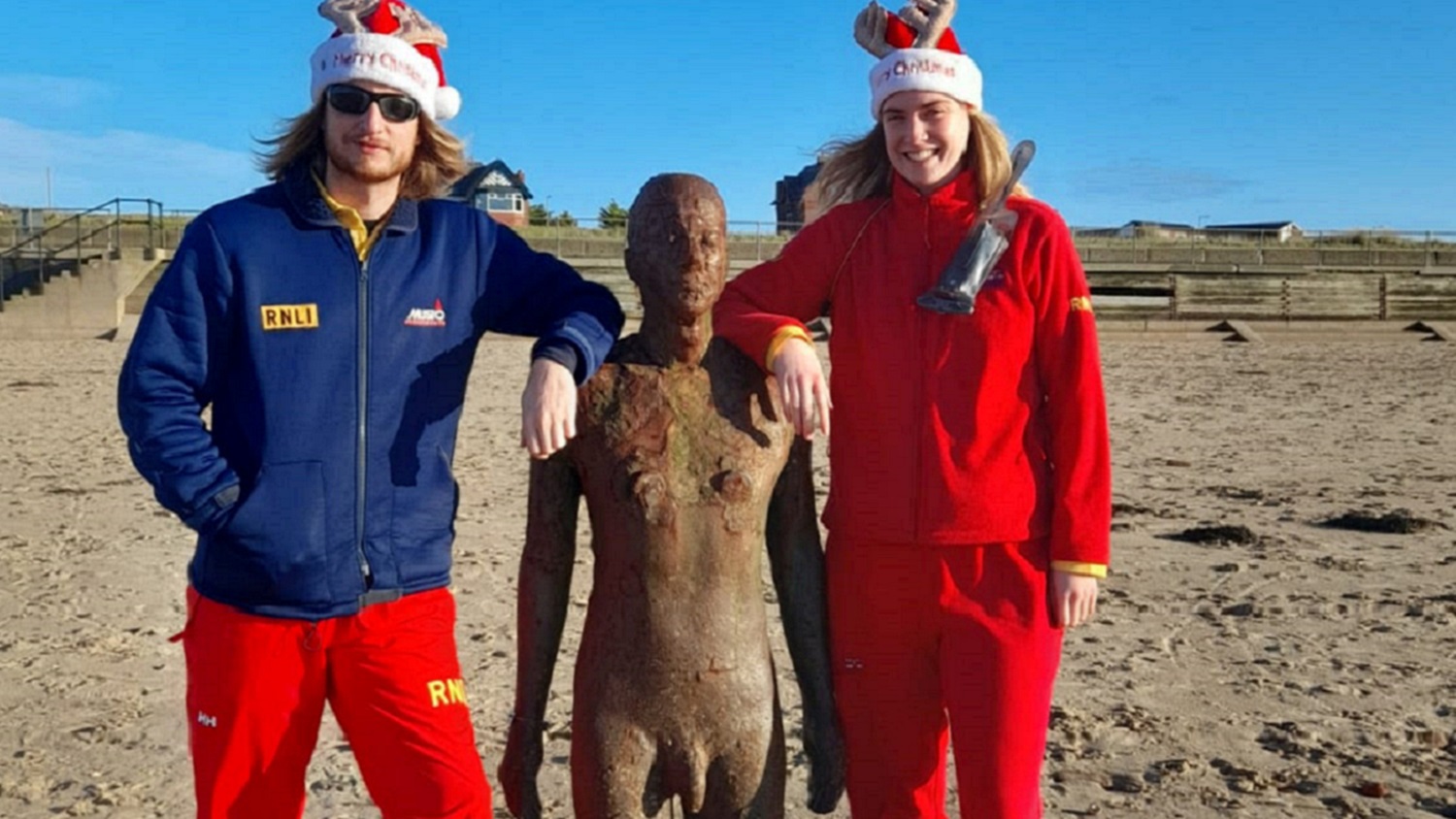 A male and female lifeguard stand with their arms around a statue on a beach in winter sun.