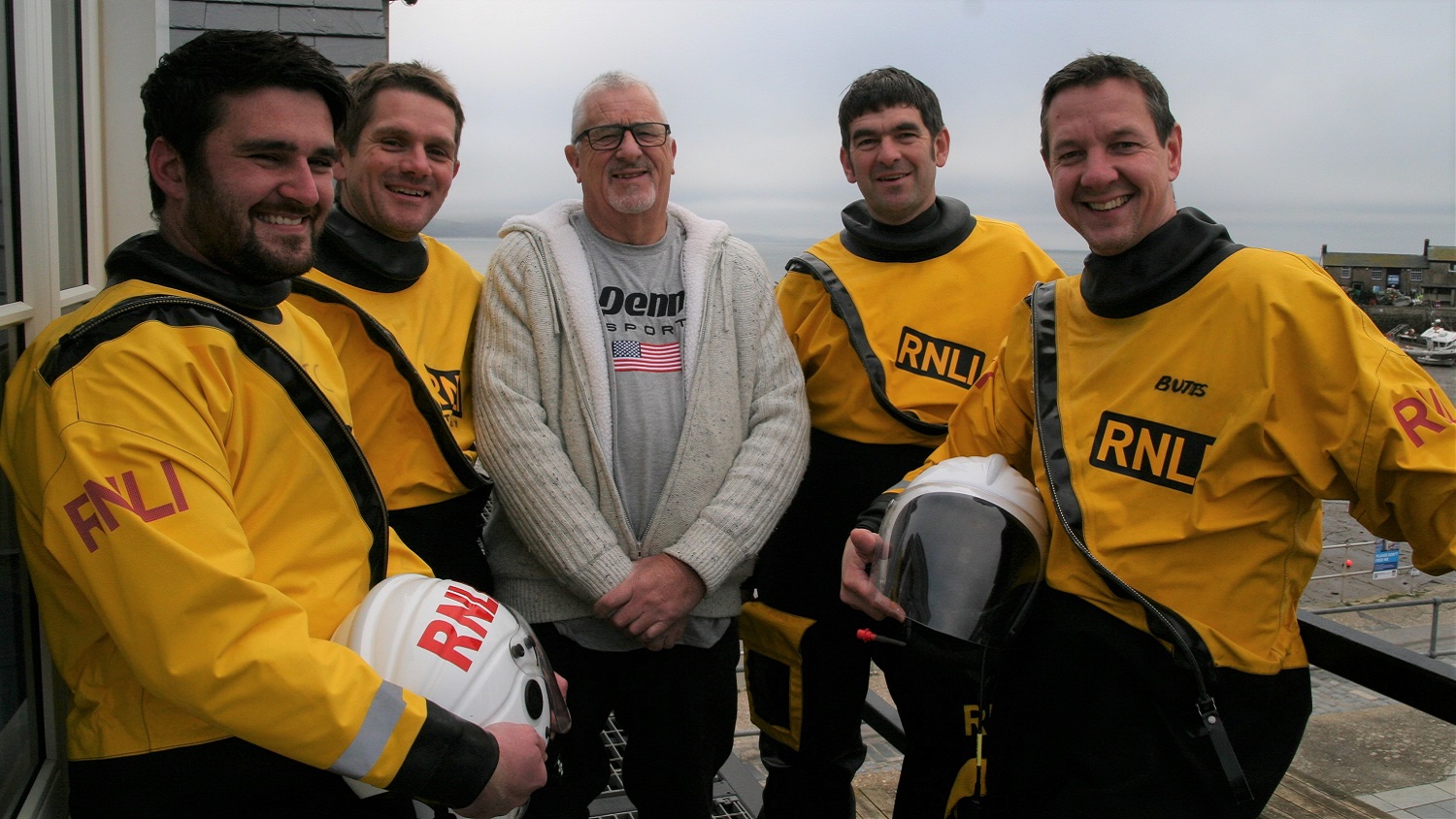 Four male RNLI crew members dressed in yellow rescue clothing stand around a middle ages man with the sea in the background.