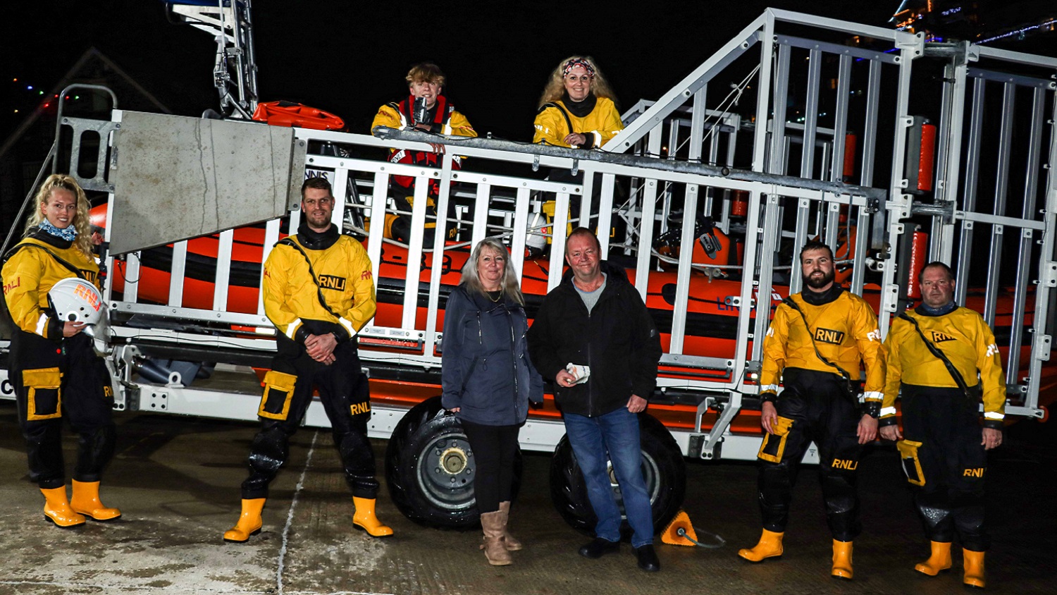 Six RNLI crew stand outside on their sea tractor in yellow lifesaving kit at night. A man and a woman stand in between the crew.