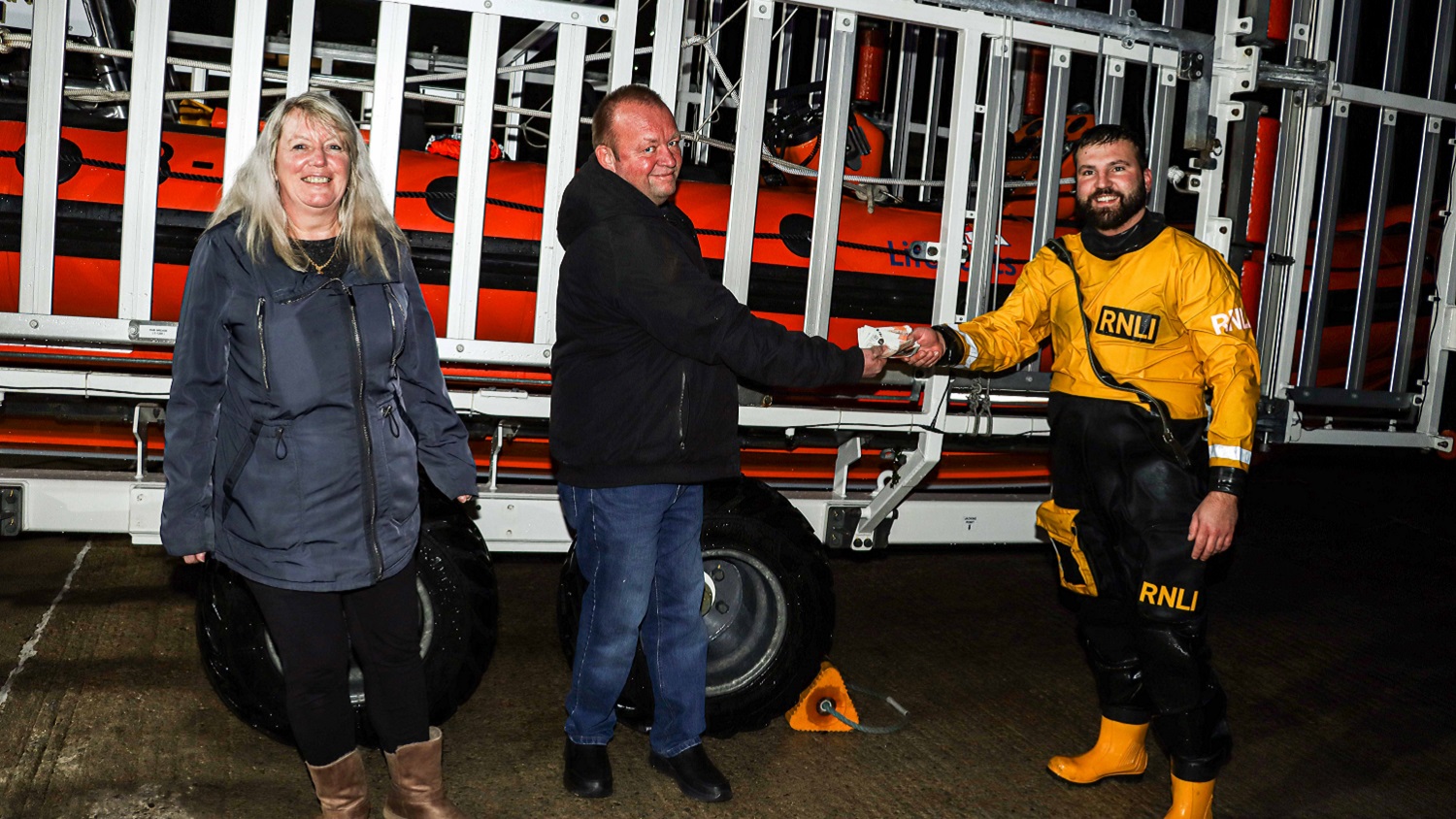 A man and woman stand in front of a sea tractor at night. The man shakes hands with a male RNLI crew member dressed in yellow rescue kit.