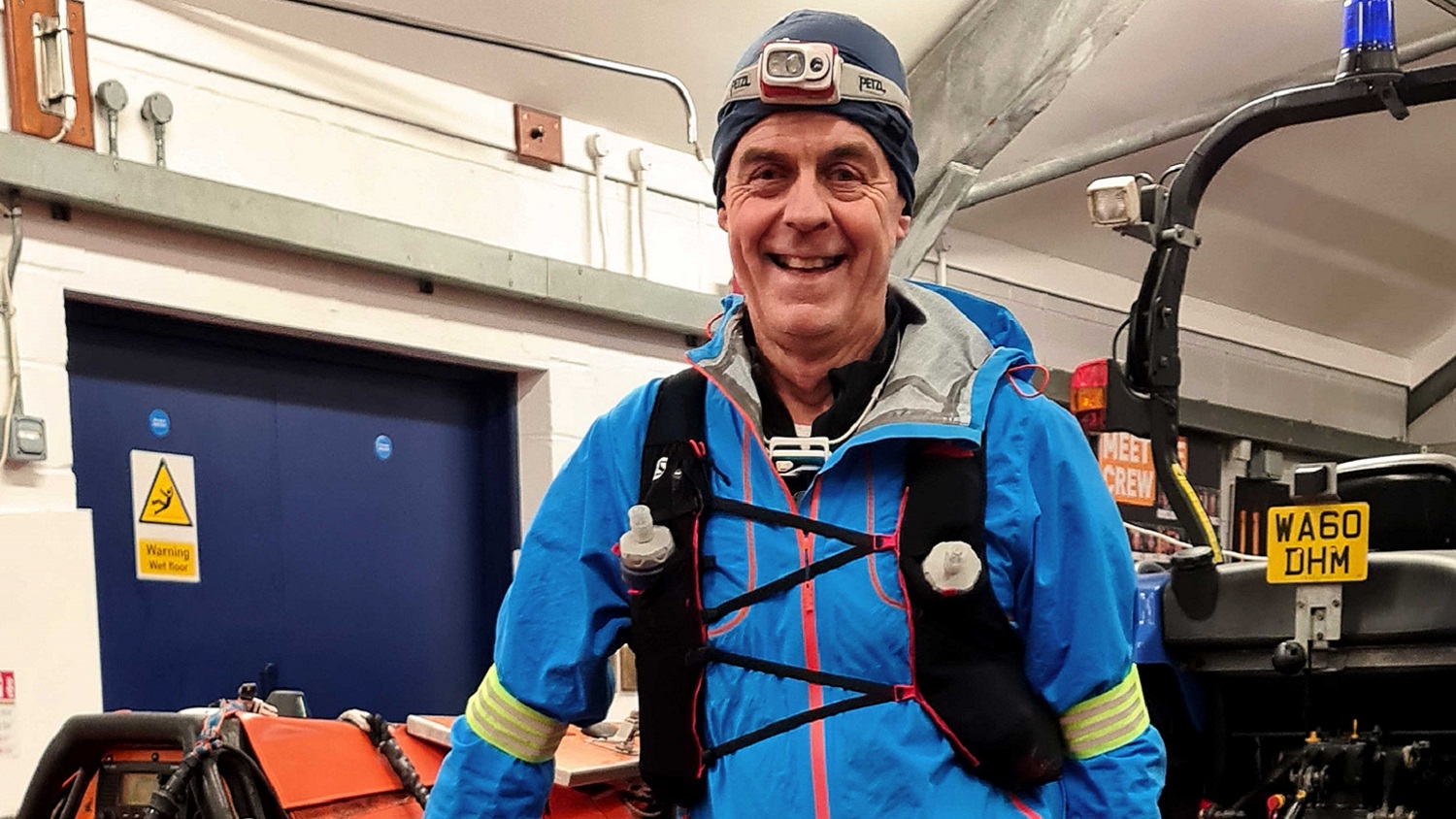 A man in winter running kit smiles broadly at the camera with an orange lifeboat visible behind him.