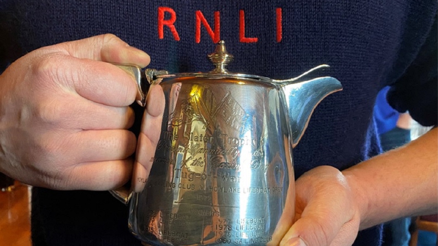 A pair of hands holds a silver commemorative teapot.