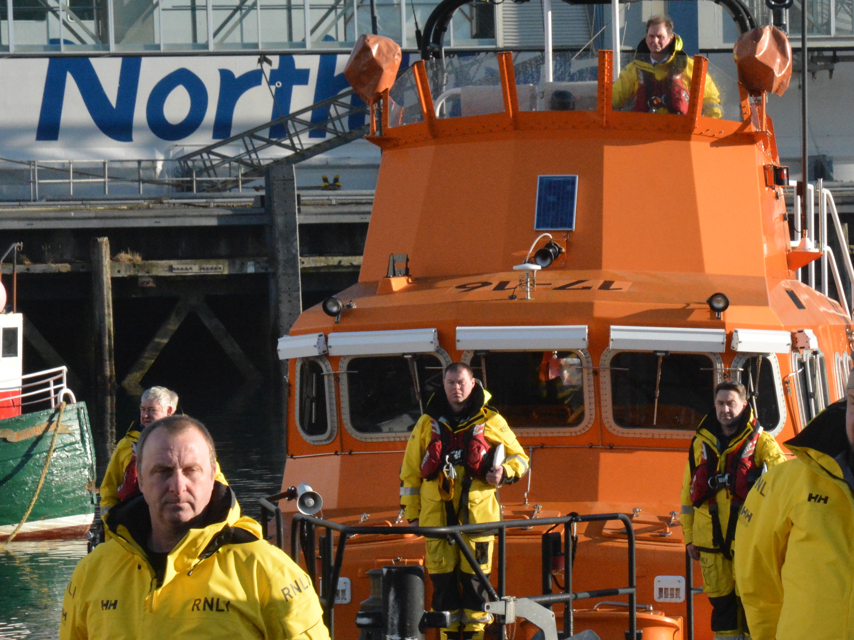Crew, caps doffed and at attention on prow of boat as hearse passes.