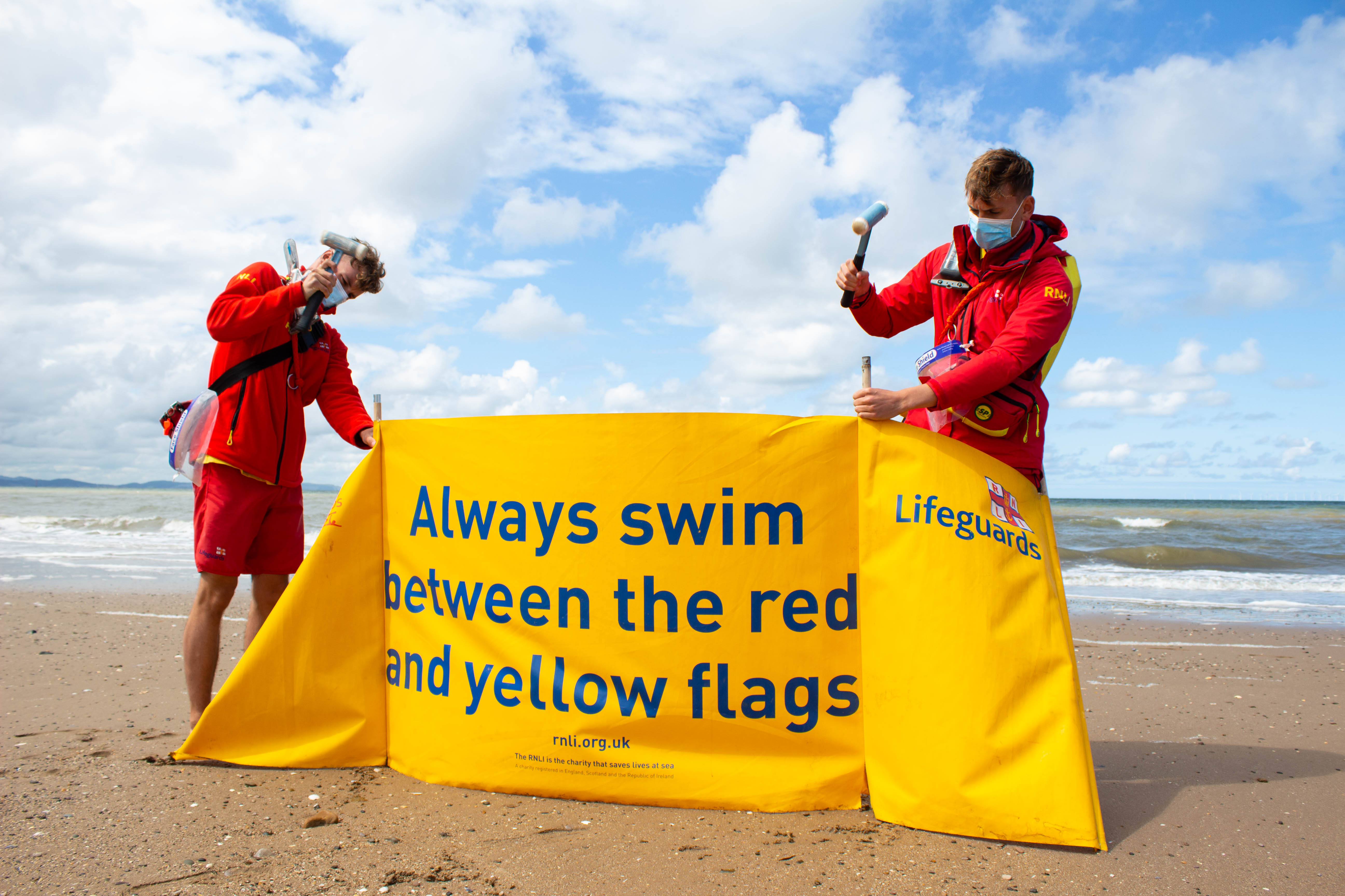 Two RNLI lifeguards putting up windbreak with safety messaging, both wearing face masks.