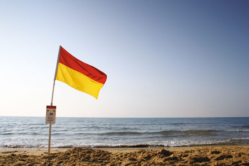 A Red and Yellow flag flies at a sandy beach. Below it, a sign tells people to swim in a lifeguarded area. Behind it, there is the sea.