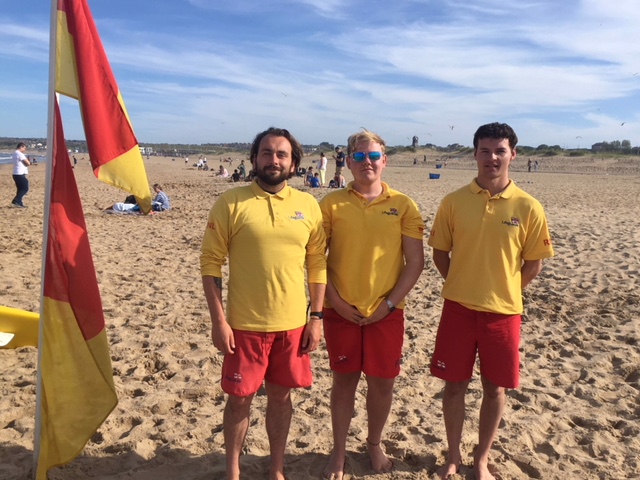Three RNLI lifeguards stand on a sandy beach. All are wearing yellow tops with RNLI on them and red shorts. Next to them are red and yellow flags.