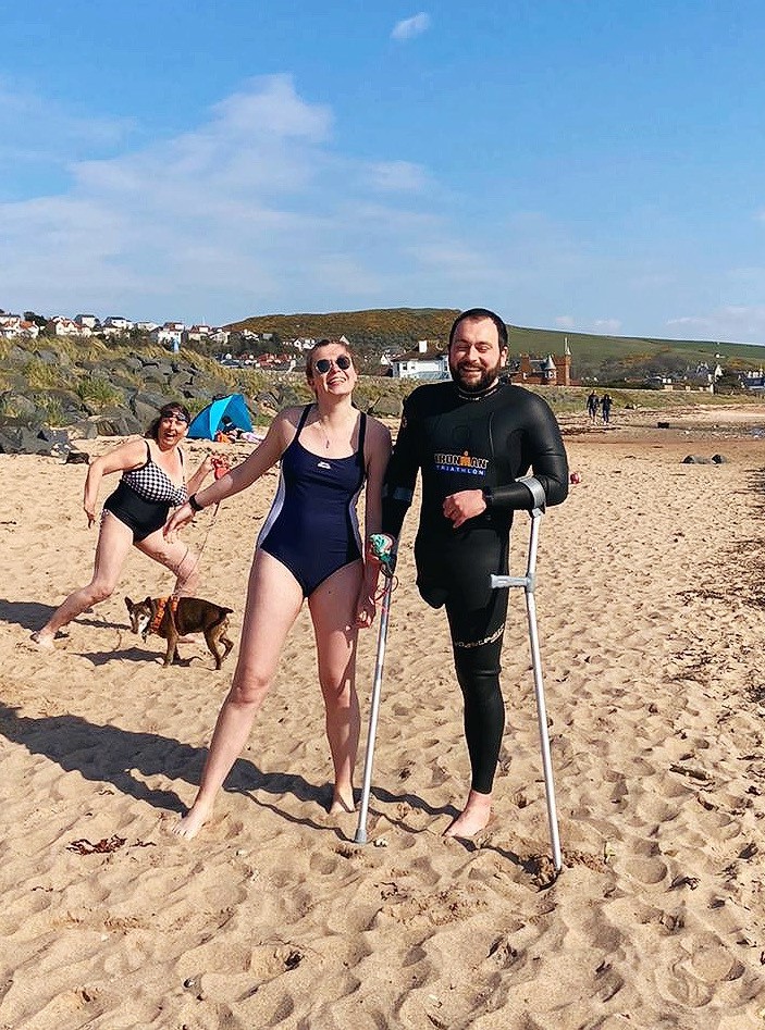A man with an amputated leg stands on the beach with crutches next to a woman in a bathing suit.