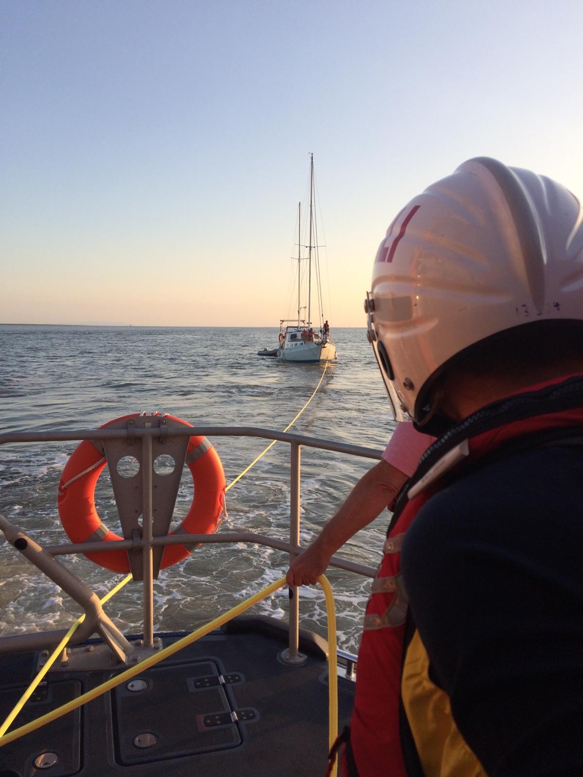 The stern of a lifeboat with a crew member and in the distance a yacht being connected by line