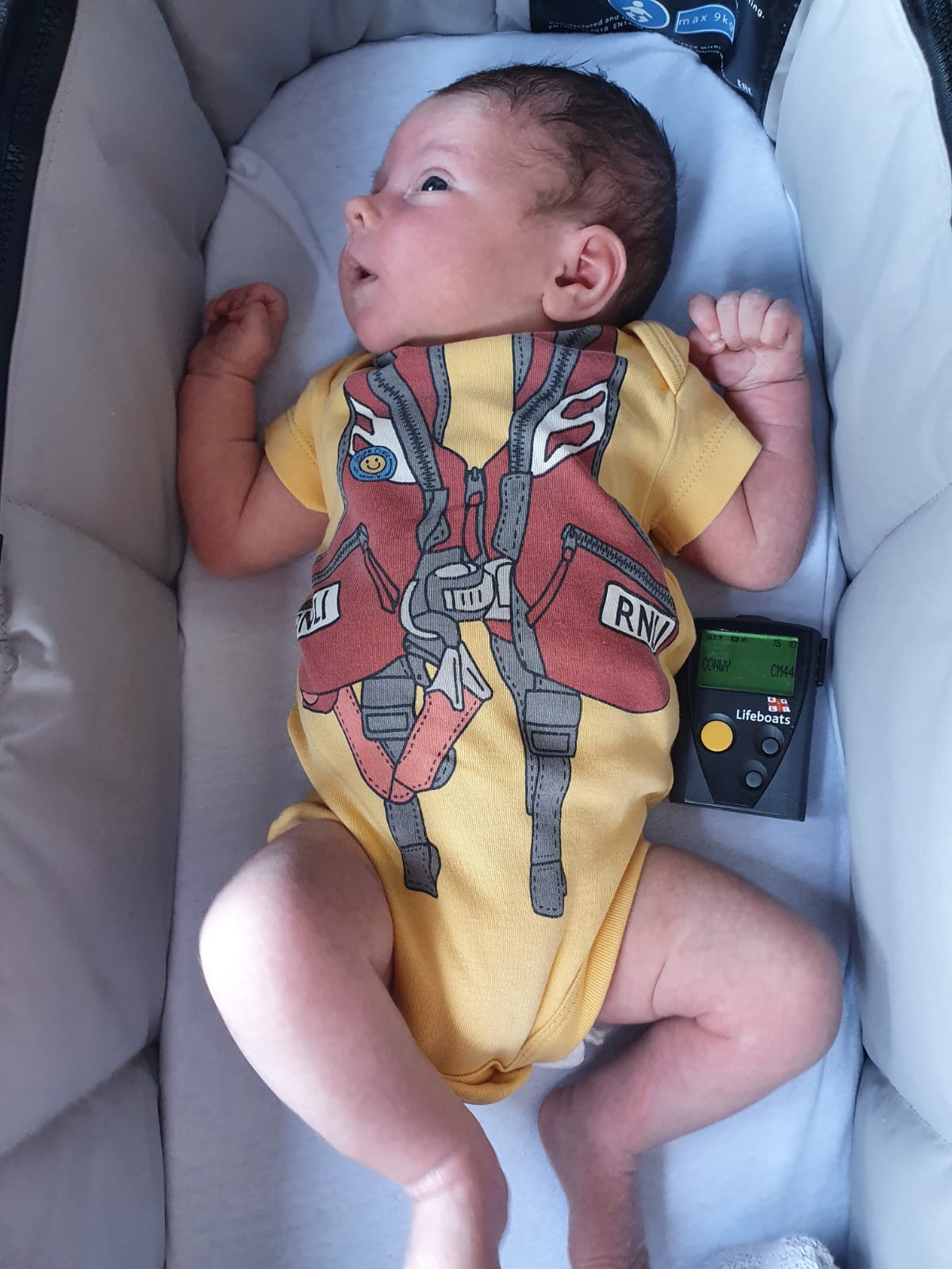Picture is Baby Harry sat in his crib alongside his Dad's pager. He is dressed in an RNLI themed babygrow.