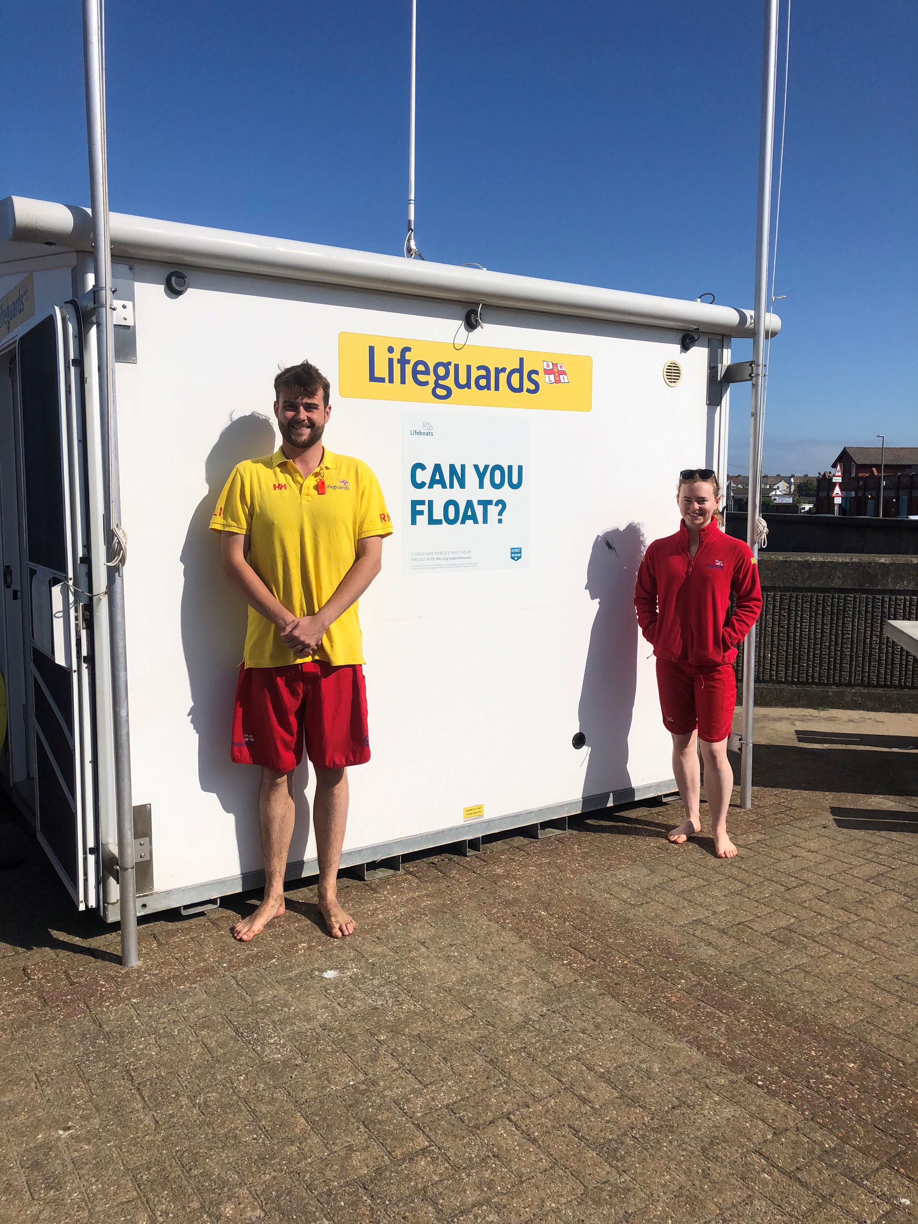 Two RNLI lifeguards in uniform stand in front of a white lifeguard hut. A yellow sign saying Lifeguards is on the hut as well as a safety message saying 'Can you float?'
