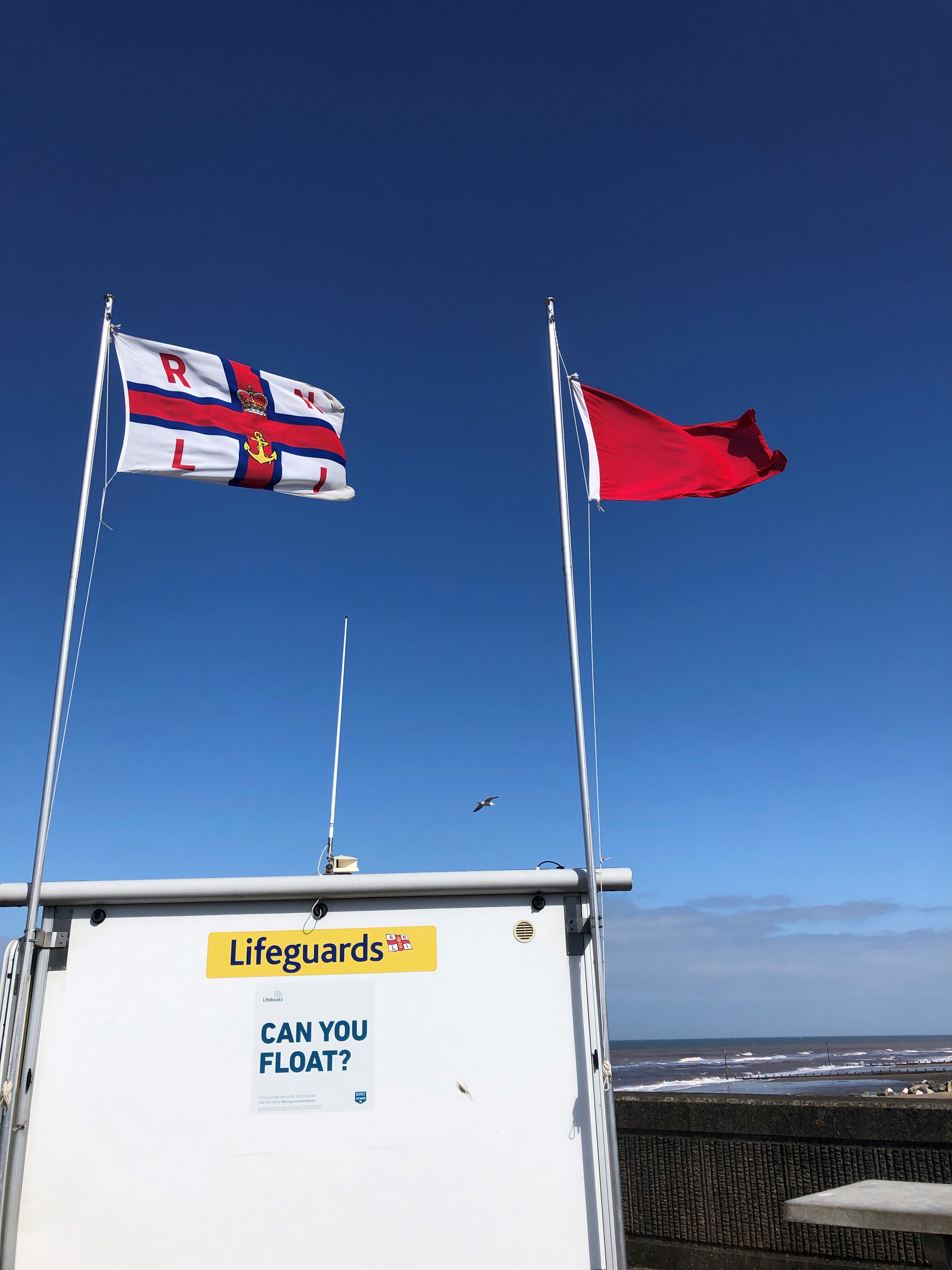 On top of a white lifeguard hut, which has a yellow sign saying Lifeguards and a safety message saying 'Can you float?', flies two flags. The flag on the left is white and has a red cross with the letters RNLI in each of the four sections. Beside it, a red flag flies meaning that people cannot swim at the beach.
