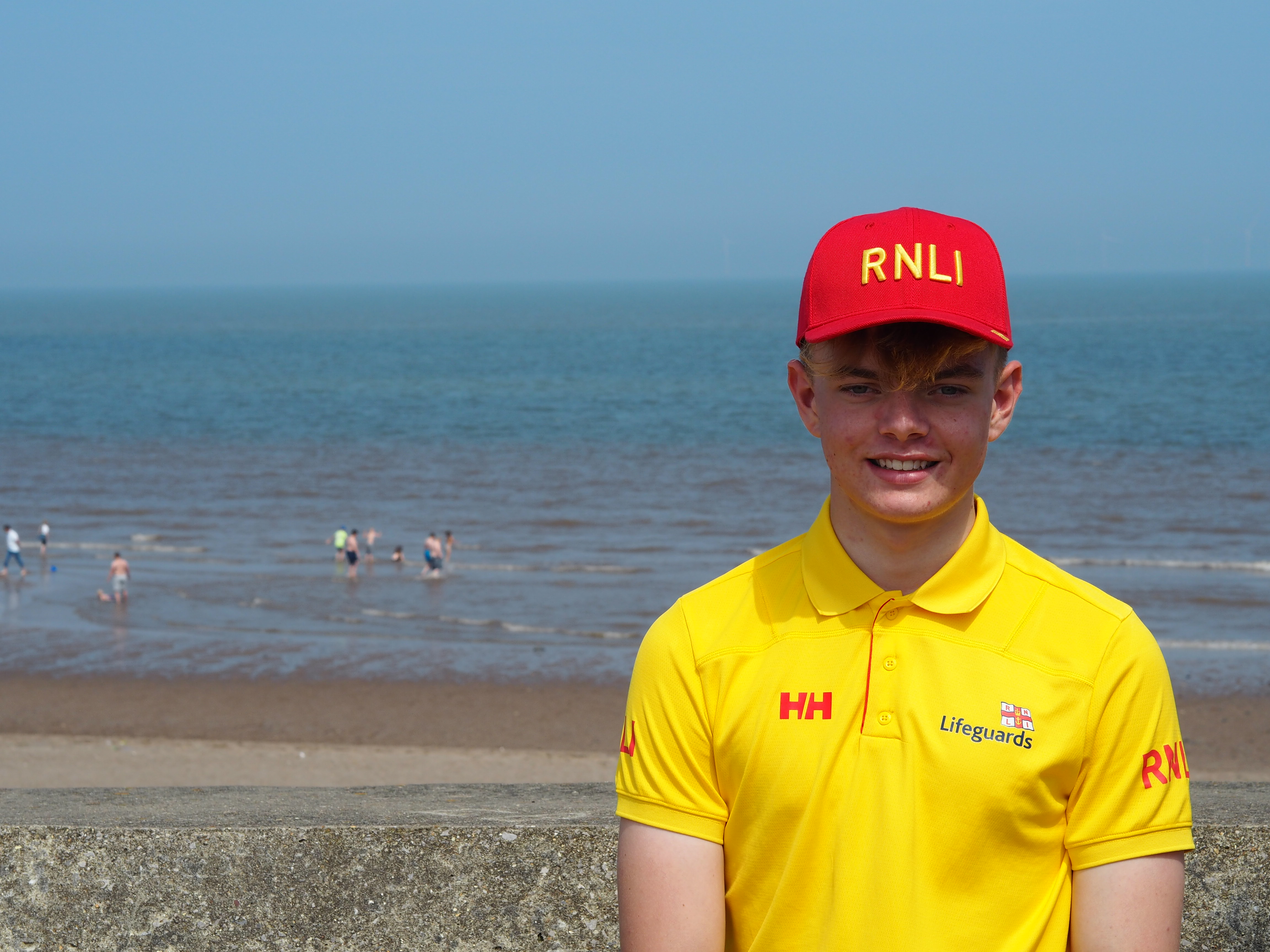 A young male in a yellow lifeguard top with a red hat that says RNLI on it stands on a rocky beach in front of the sea.