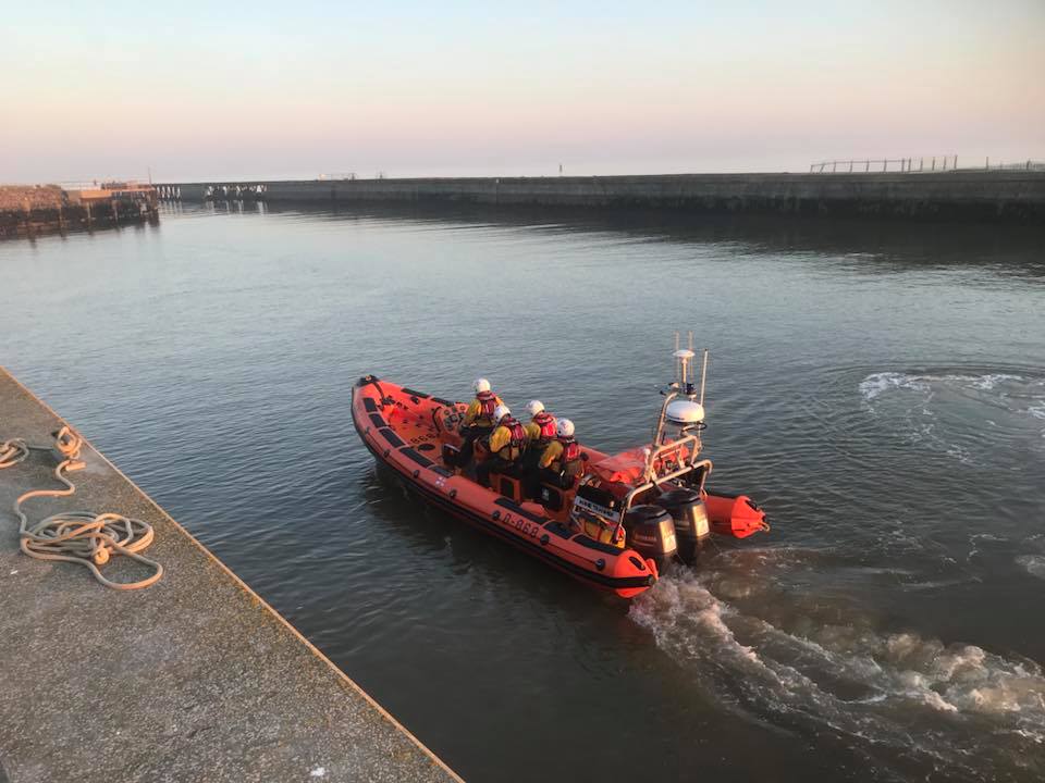 In a harbour or docks, an orange lifeboat with 3 people on board sits on the water. It is relatively small.