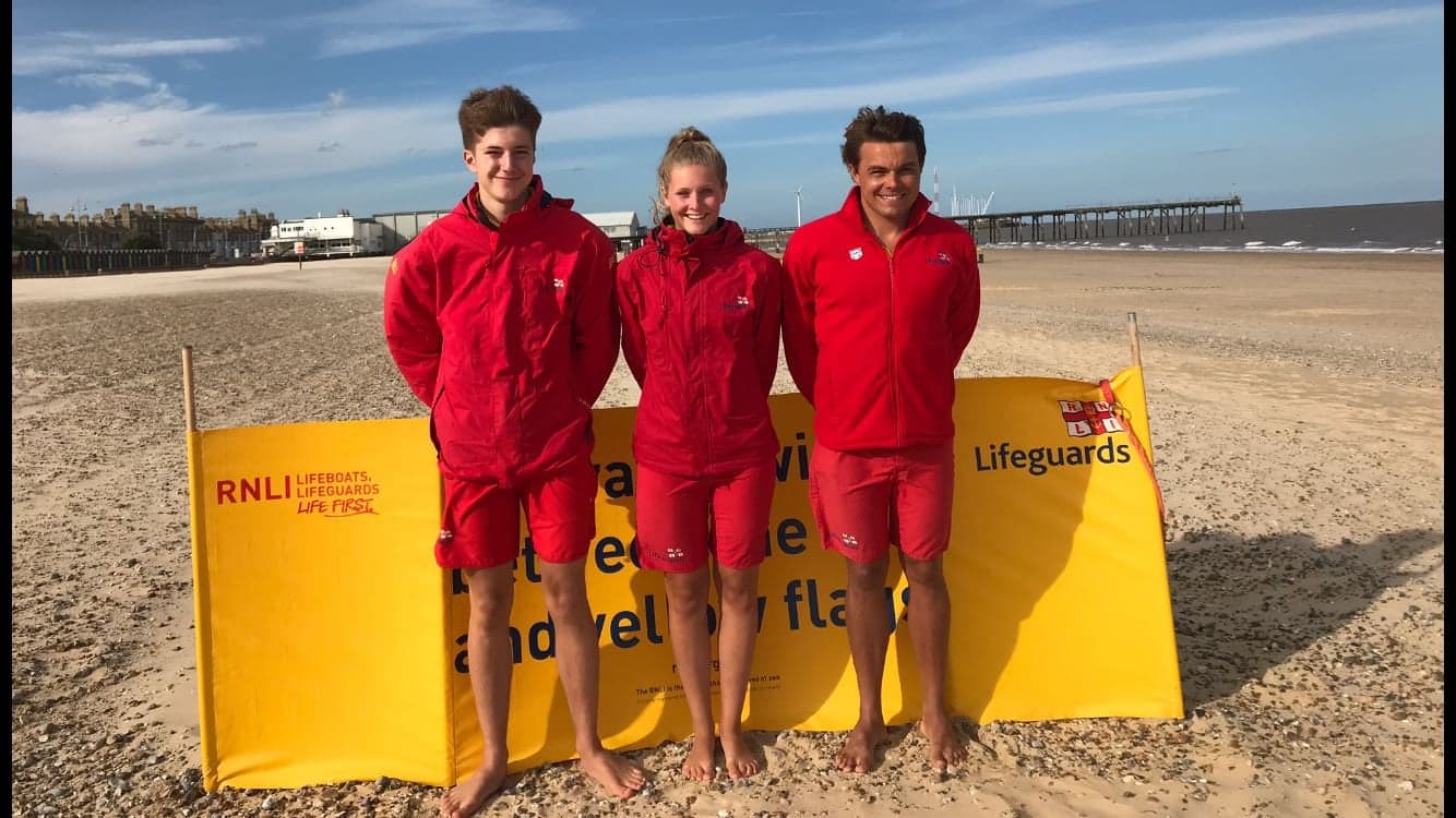 Three RNLI Lifeguards in red lifeguard kit standing in front of a yellow Lifeguard sign on a beach