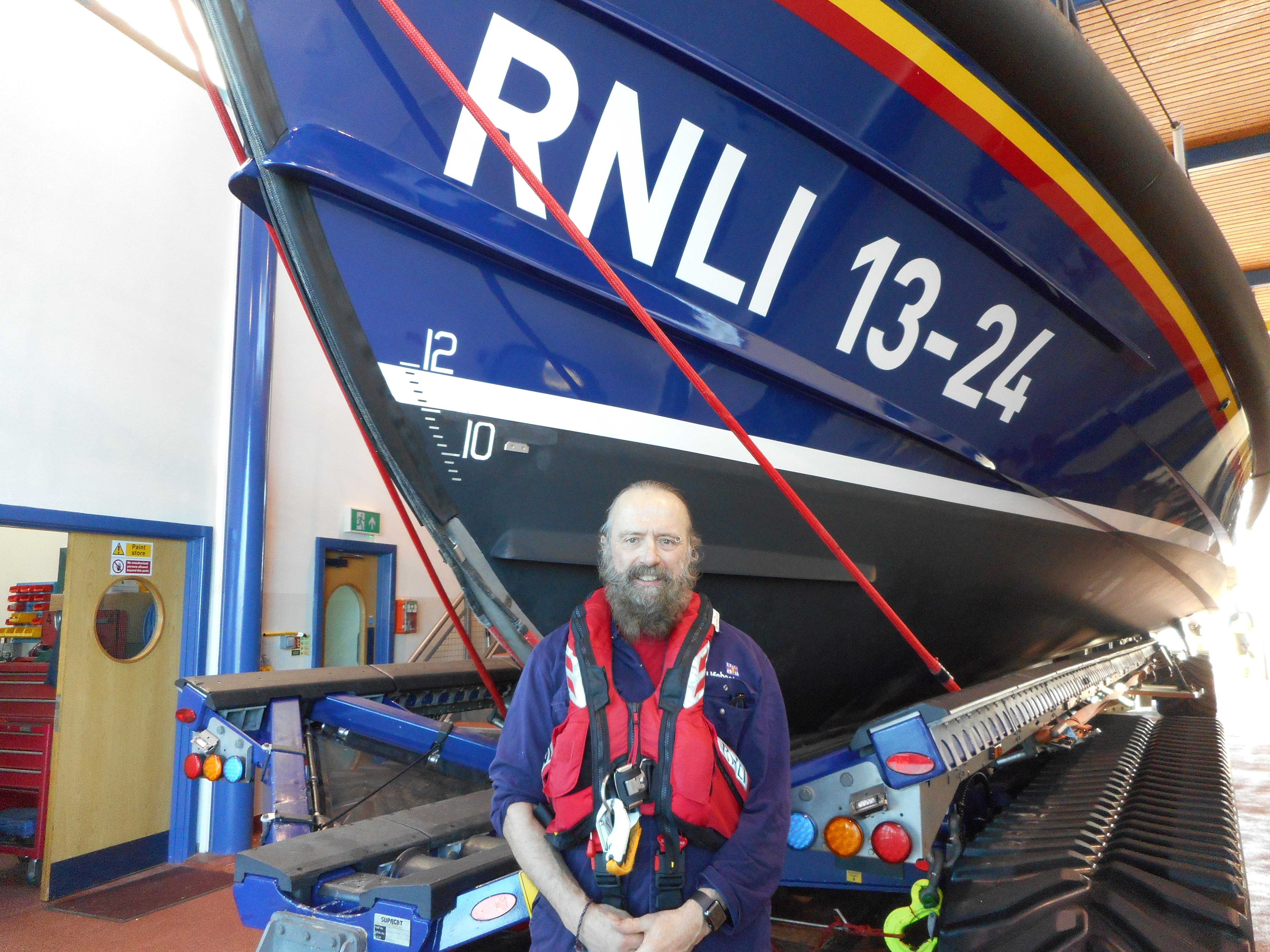 A man wearing a life jacket stands in front of a lifeboat 