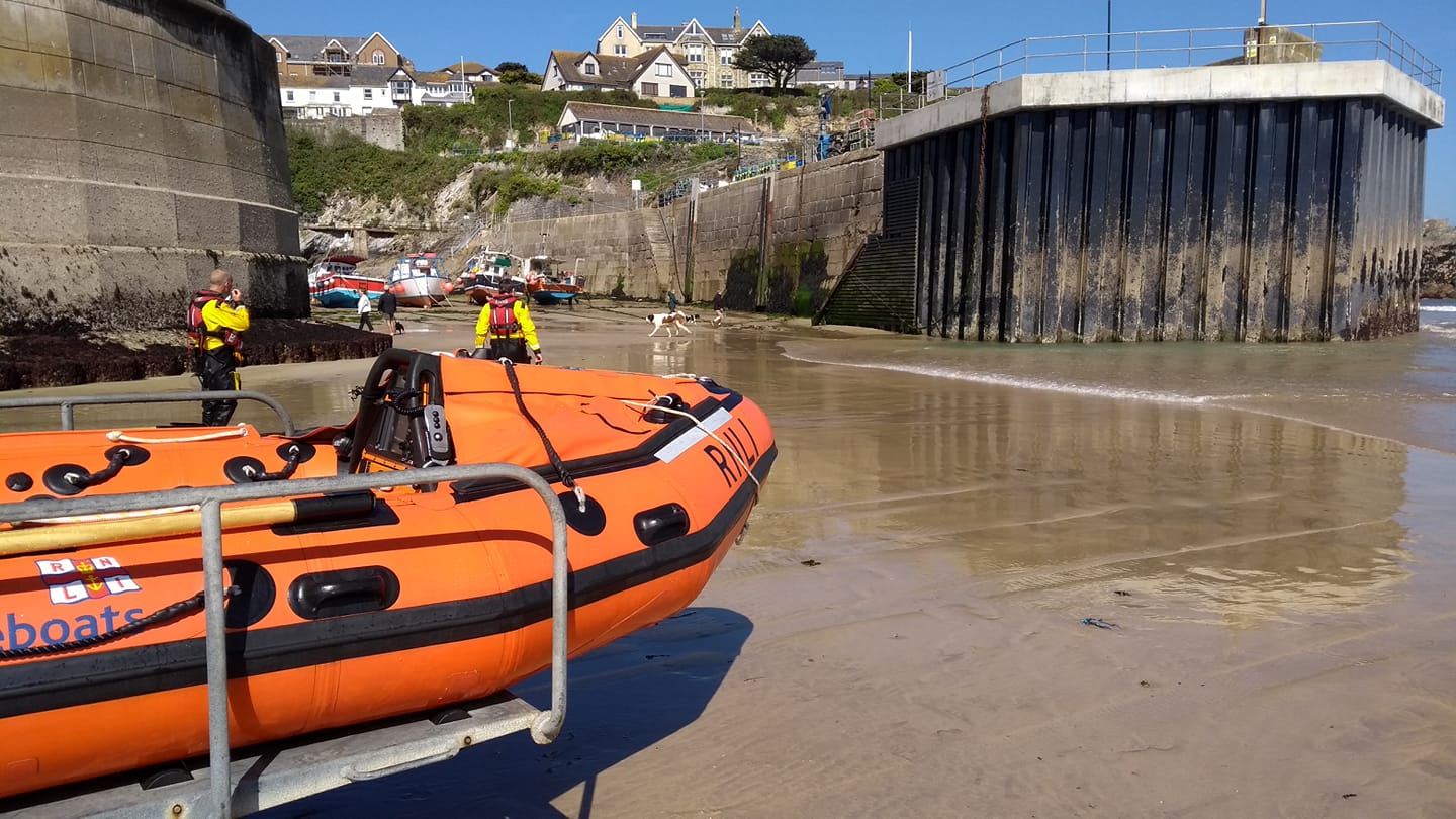 Newquay RNLI D class inshore lifeboat on the beach in the foreground, with RNLI crew walking through the harbour entrance at low tide
