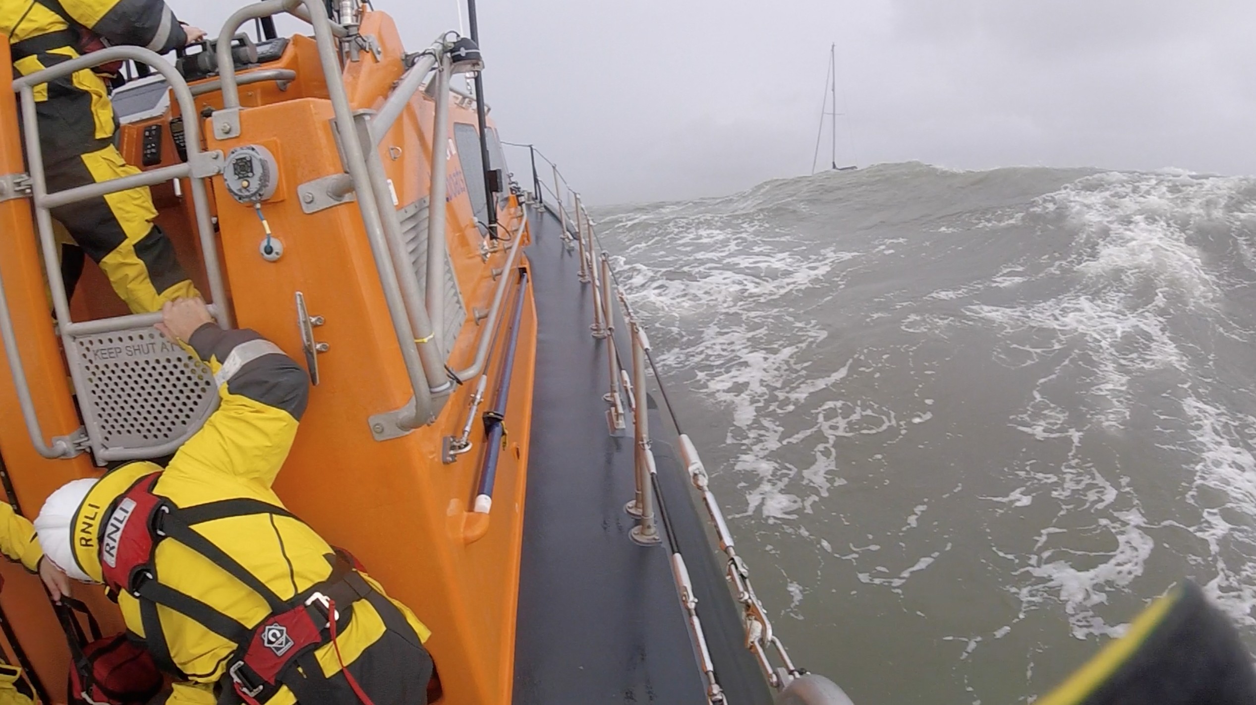 a lifeboat is seen with just the mast of a yacht appearing over a large wave