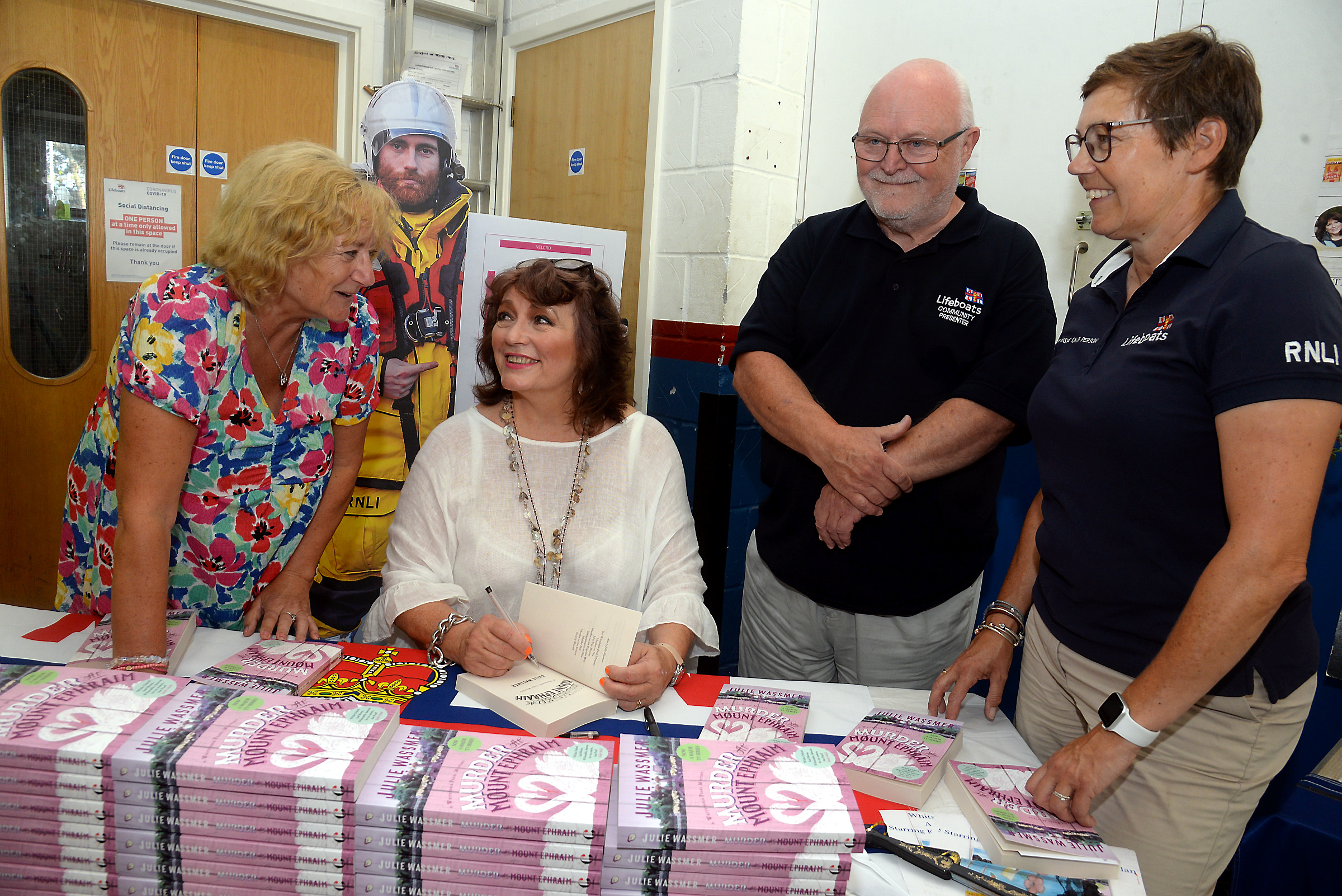 Whitstable author Julie Wassmer signs a copy of her latest book 'Murder at Mount Ephraim' for Keren Marwood watched by RNLI Community Presenter Gerry Skinner and Kellie Gray, Chair of Fundraising at Whitstable lifeboat station.